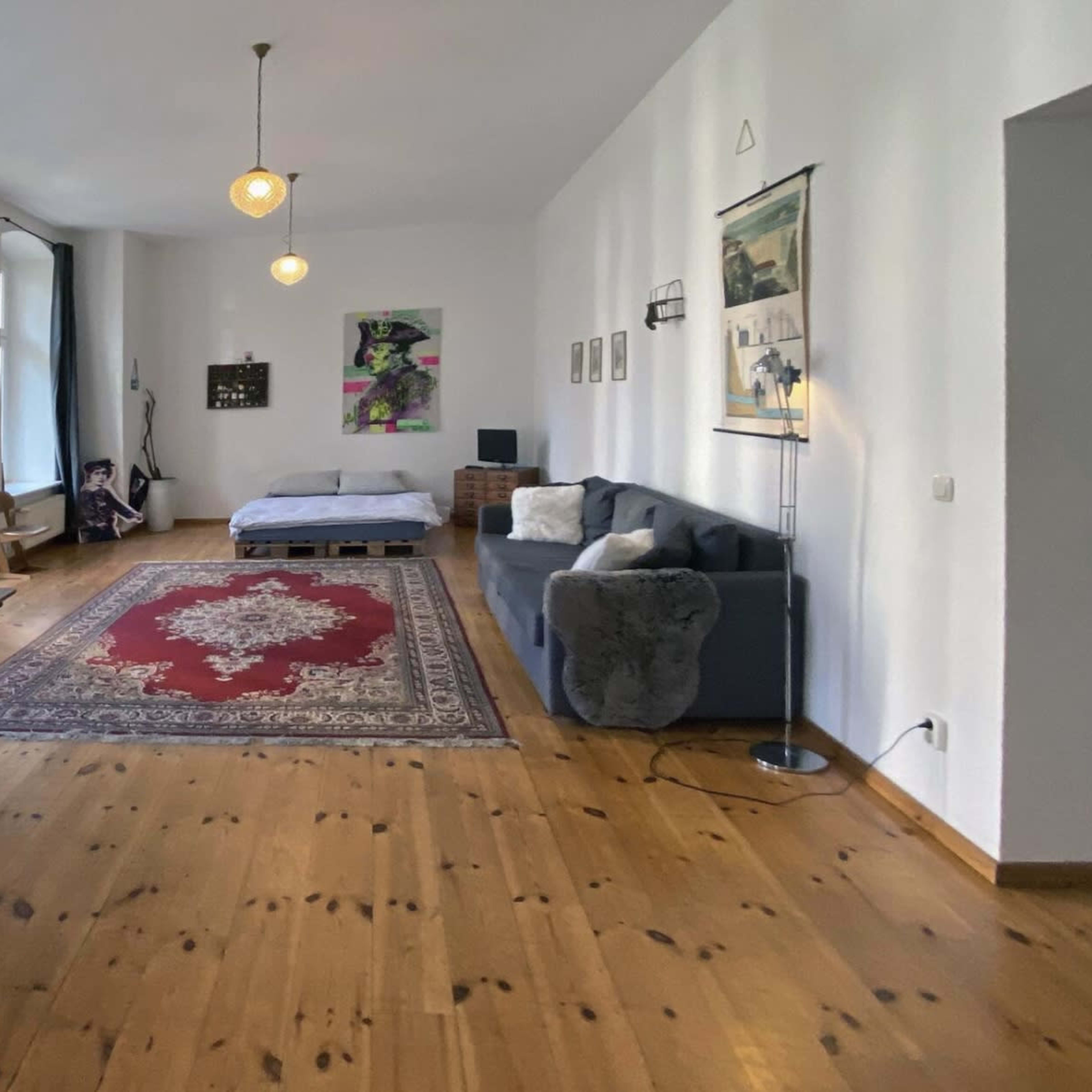 A spacious room with wooden floors, a large area rug, a gray sofa, a bed in the background, and a dining table with chairs.