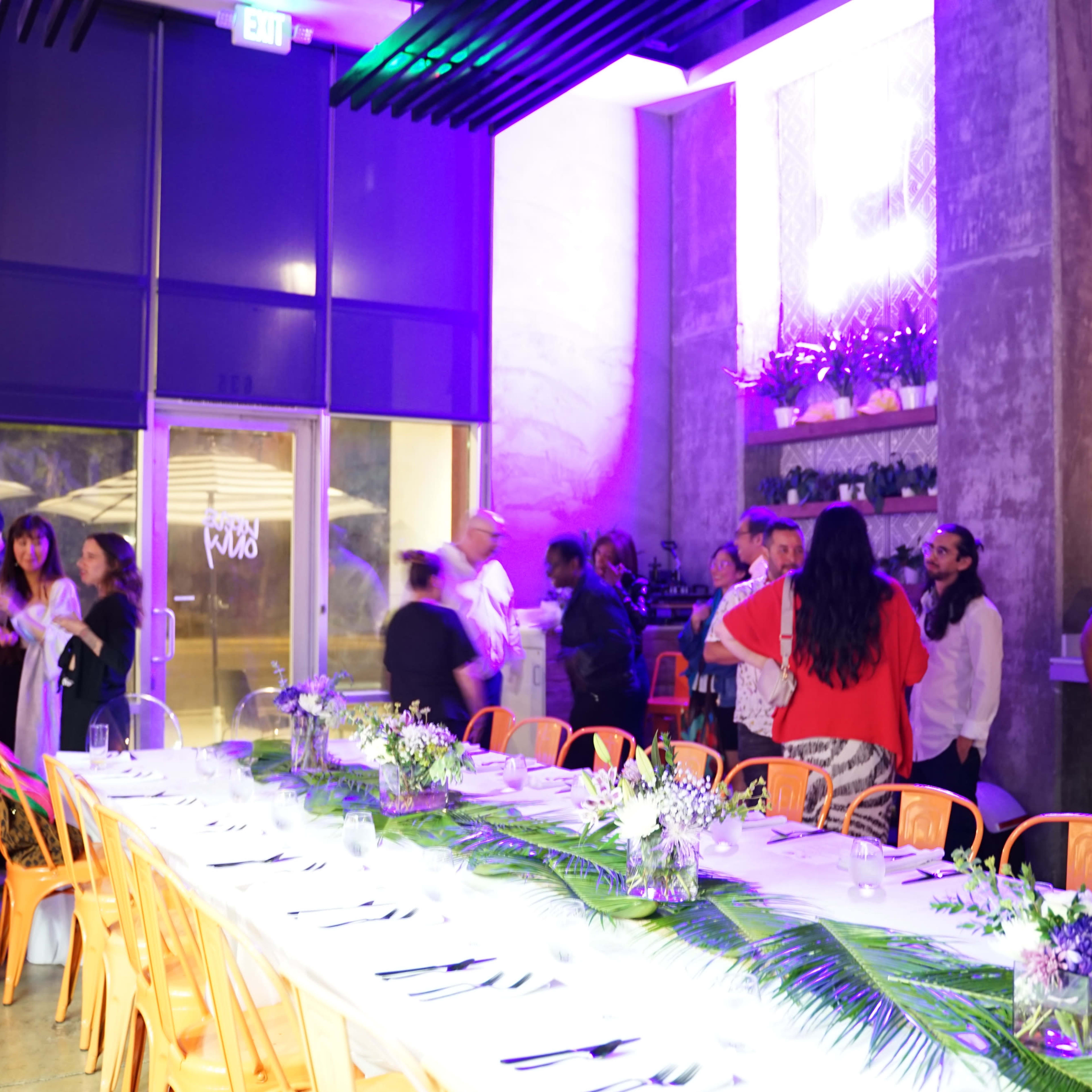 A long table is set for a gathering in a modern venue with vibrant lighting, while people socialize in small groups.