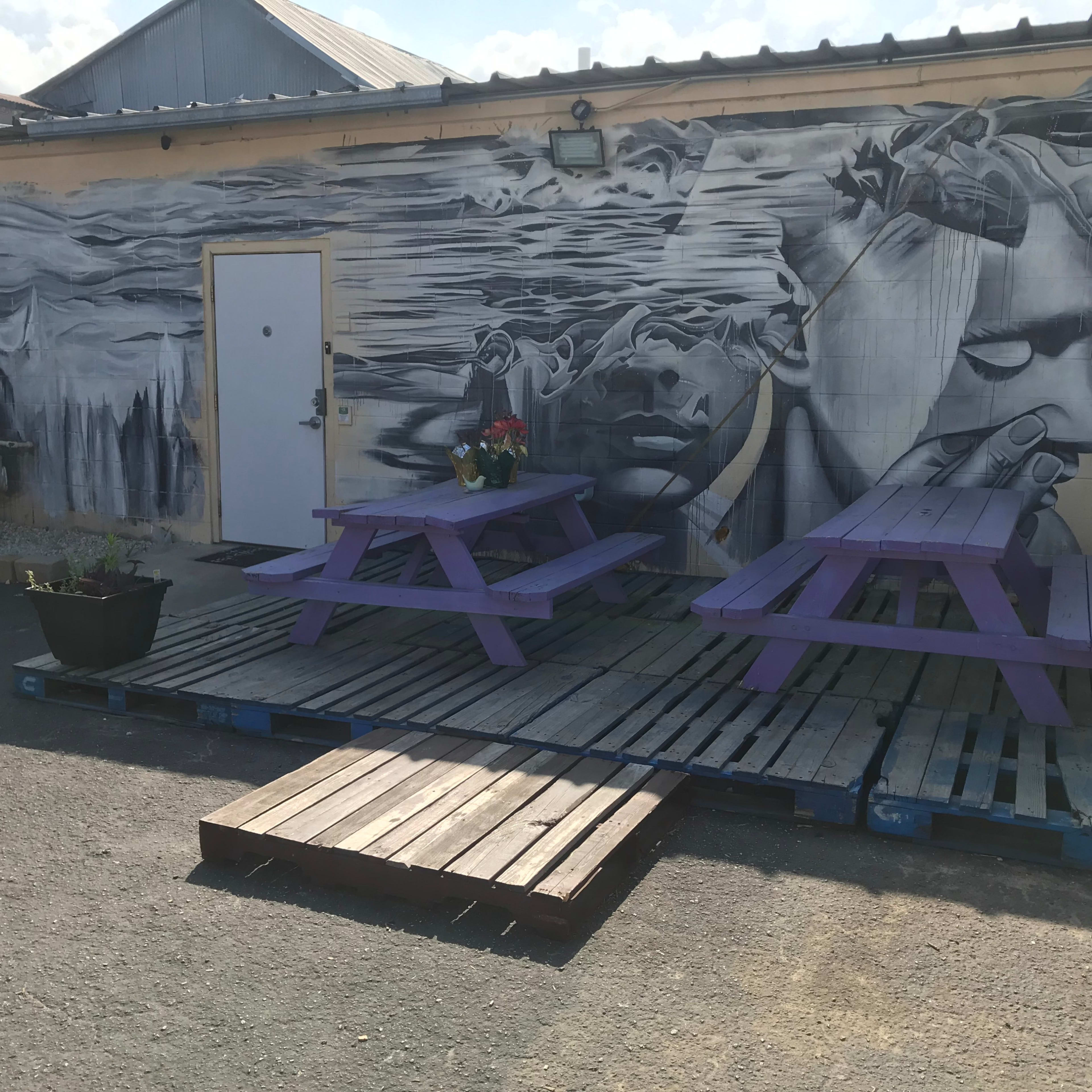 A mural featuring abstract figures on a wall beside two purple picnic tables arranged on wooden pallets in an outdoor area.