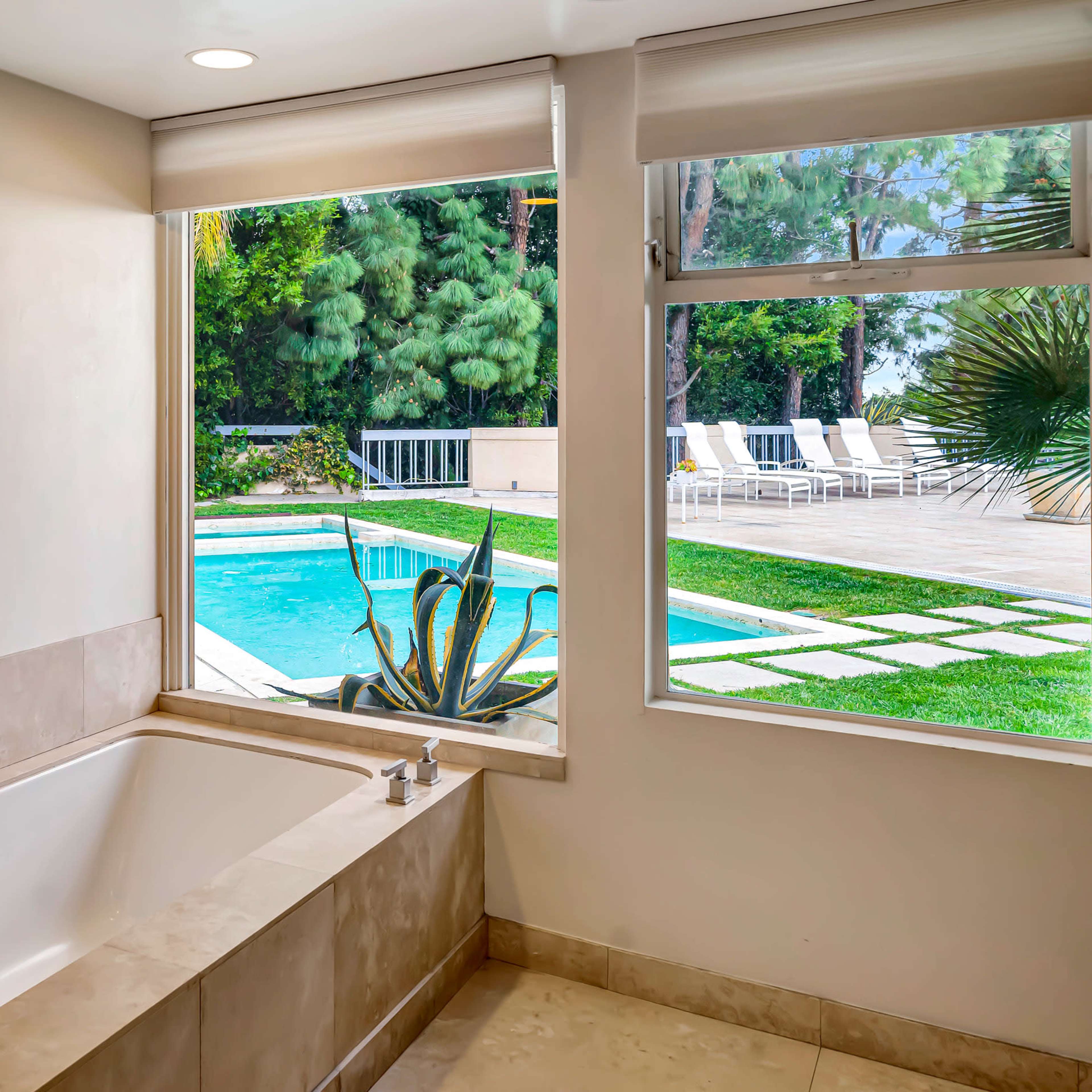 A modern bathroom features a soaking tub beside a window that overlooks a pool area and a landscaped yard.
