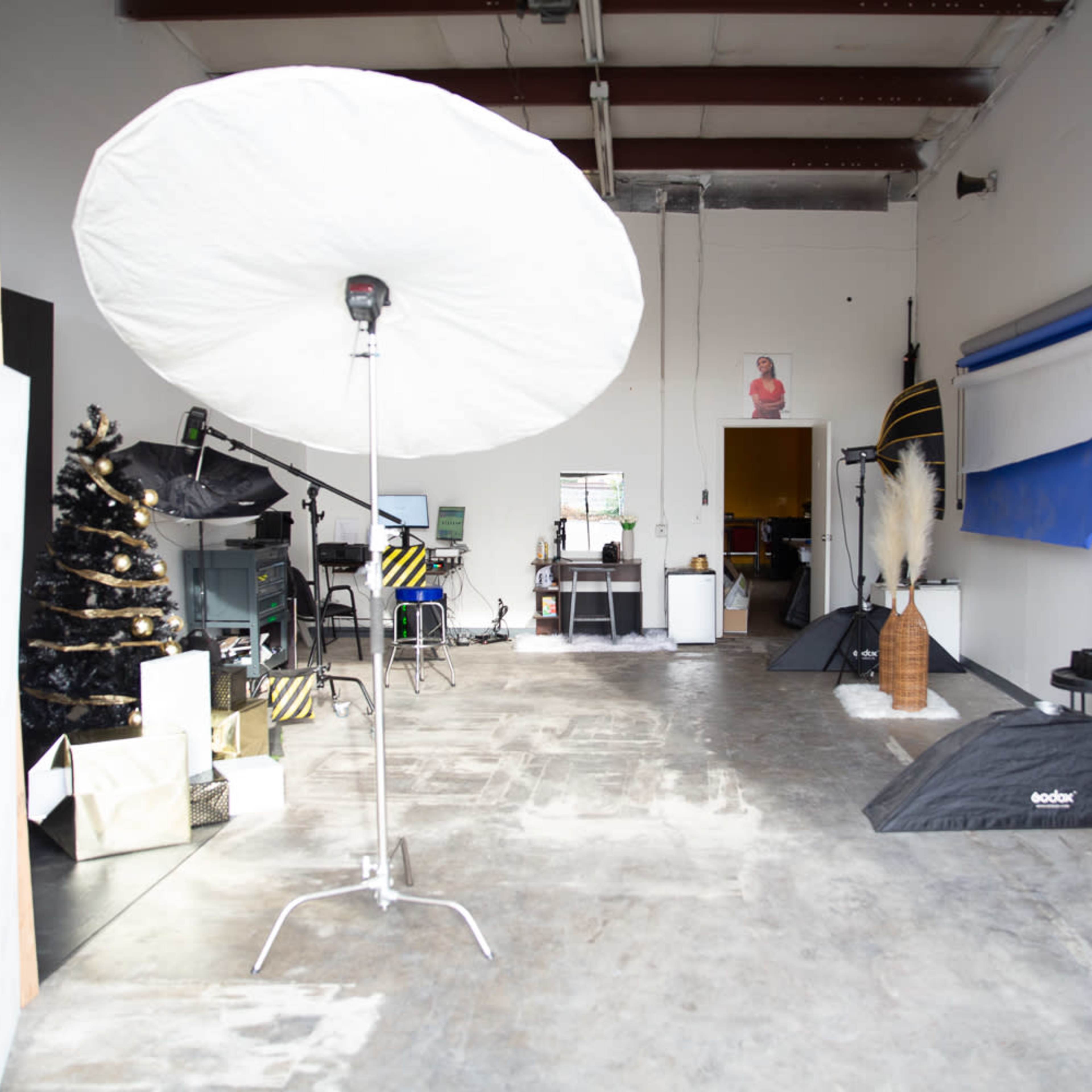A photography studio featuring a large umbrella light, a black Christmas tree, and various props and backdrops arranged throughout the space.