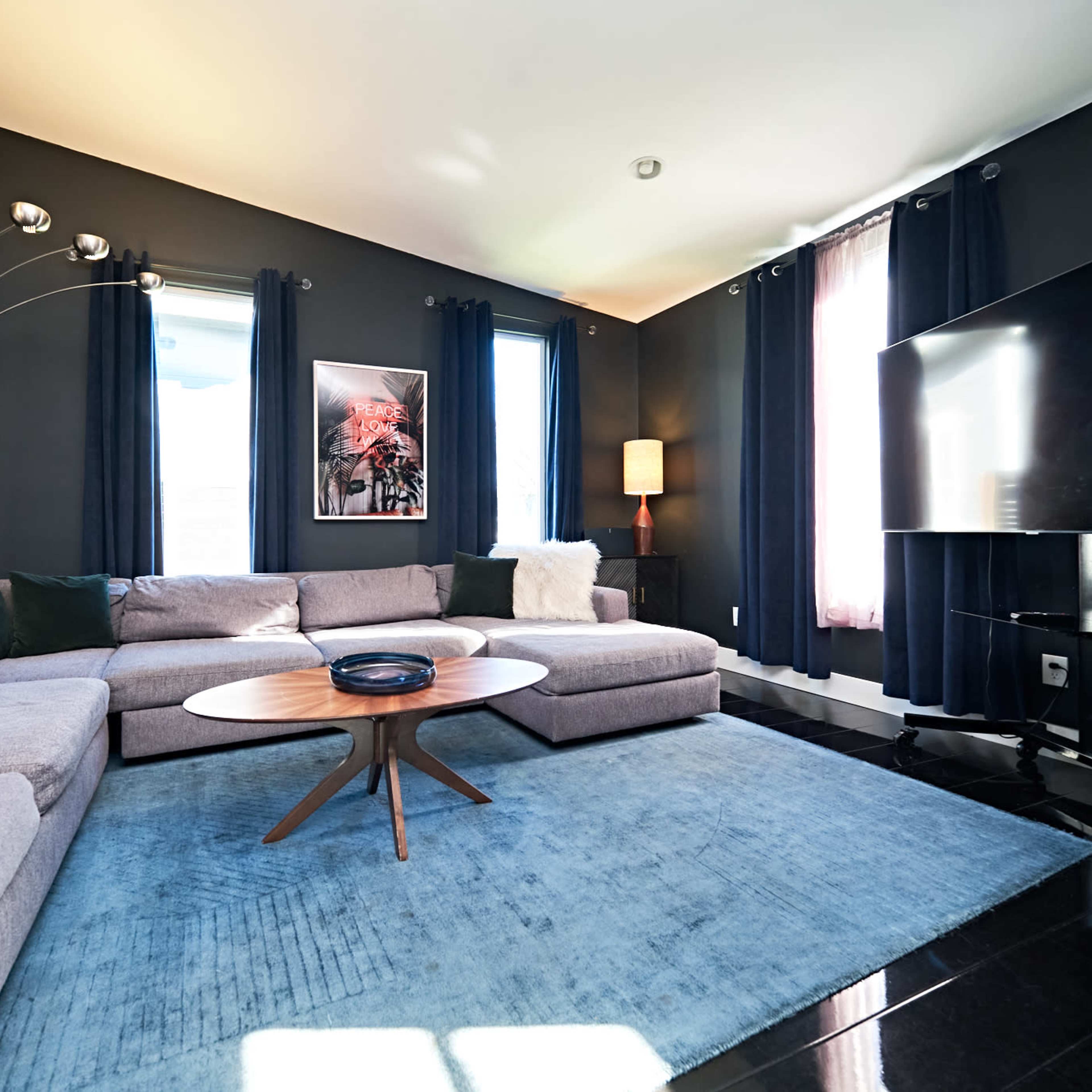 The living room features a large sectional sofa, a circular coffee table, black curtains, and a wall-mounted television against dark walls.