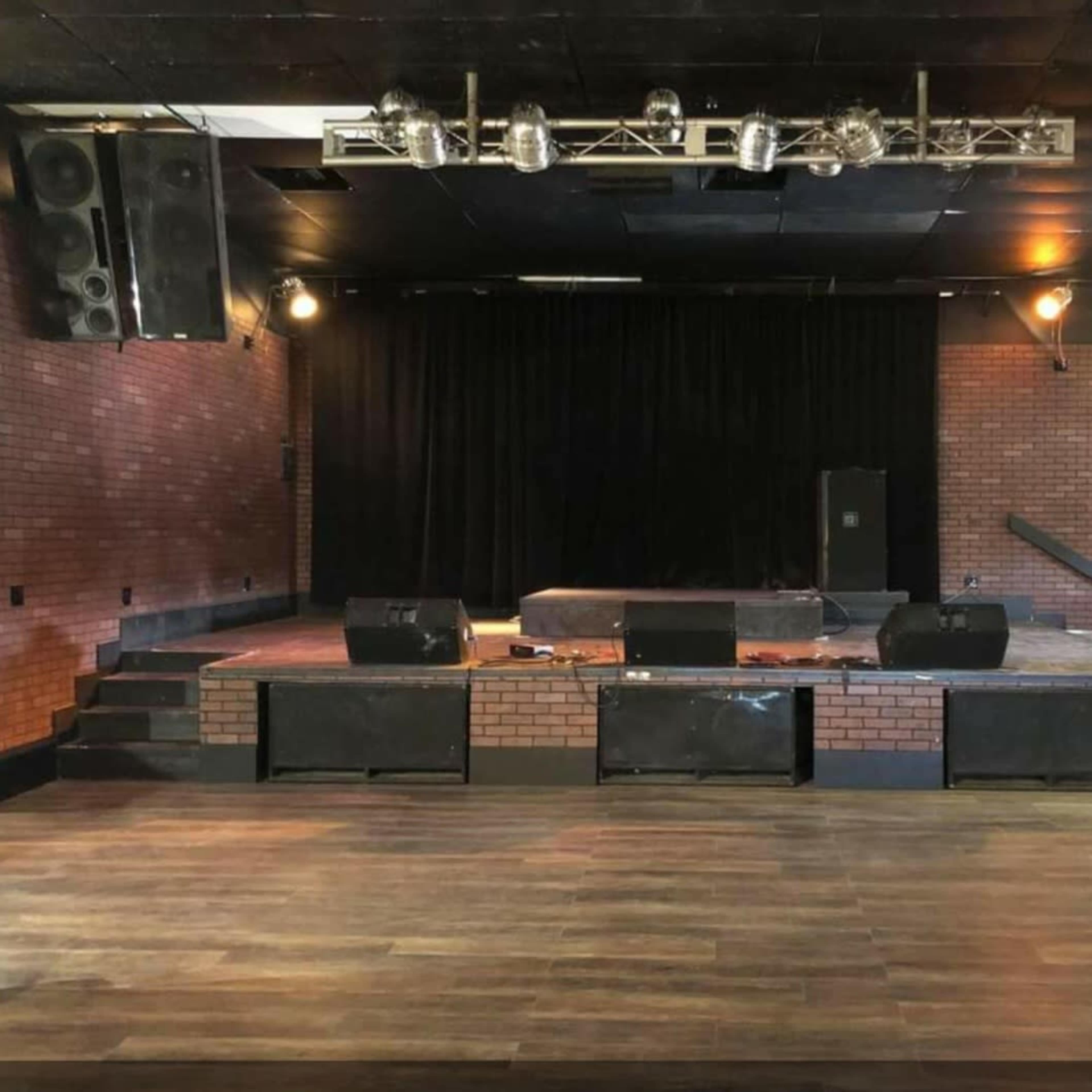 A stage area in a venue with a brick wall backdrop, wooden flooring, and sound equipment set up.