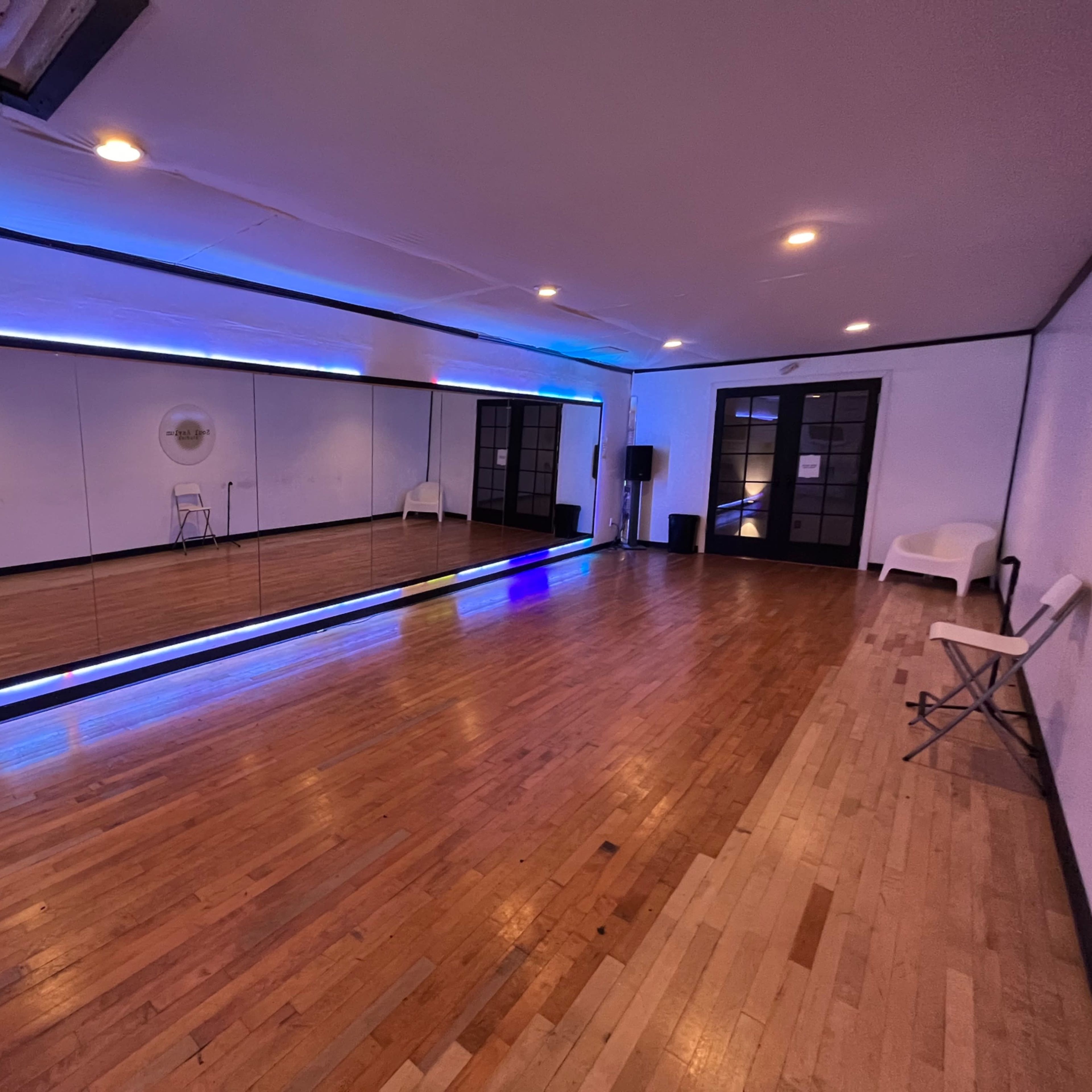 A spacious room with wooden floors, a large mirror framed by neon lights, and minimal seating along the walls.