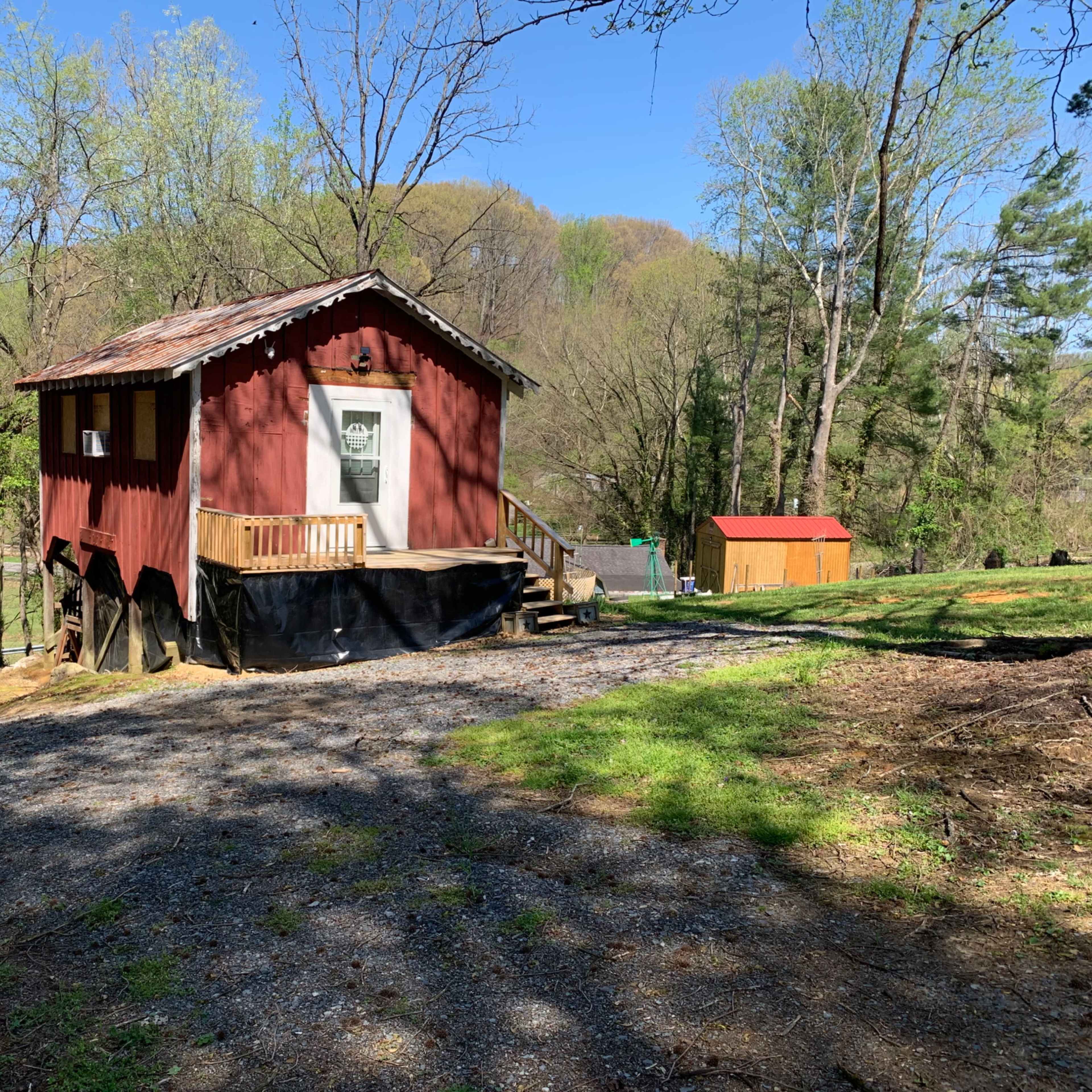 A small red house is situated on a gravel path surrounded by trees, with a covered porch and a storage shed in the background.