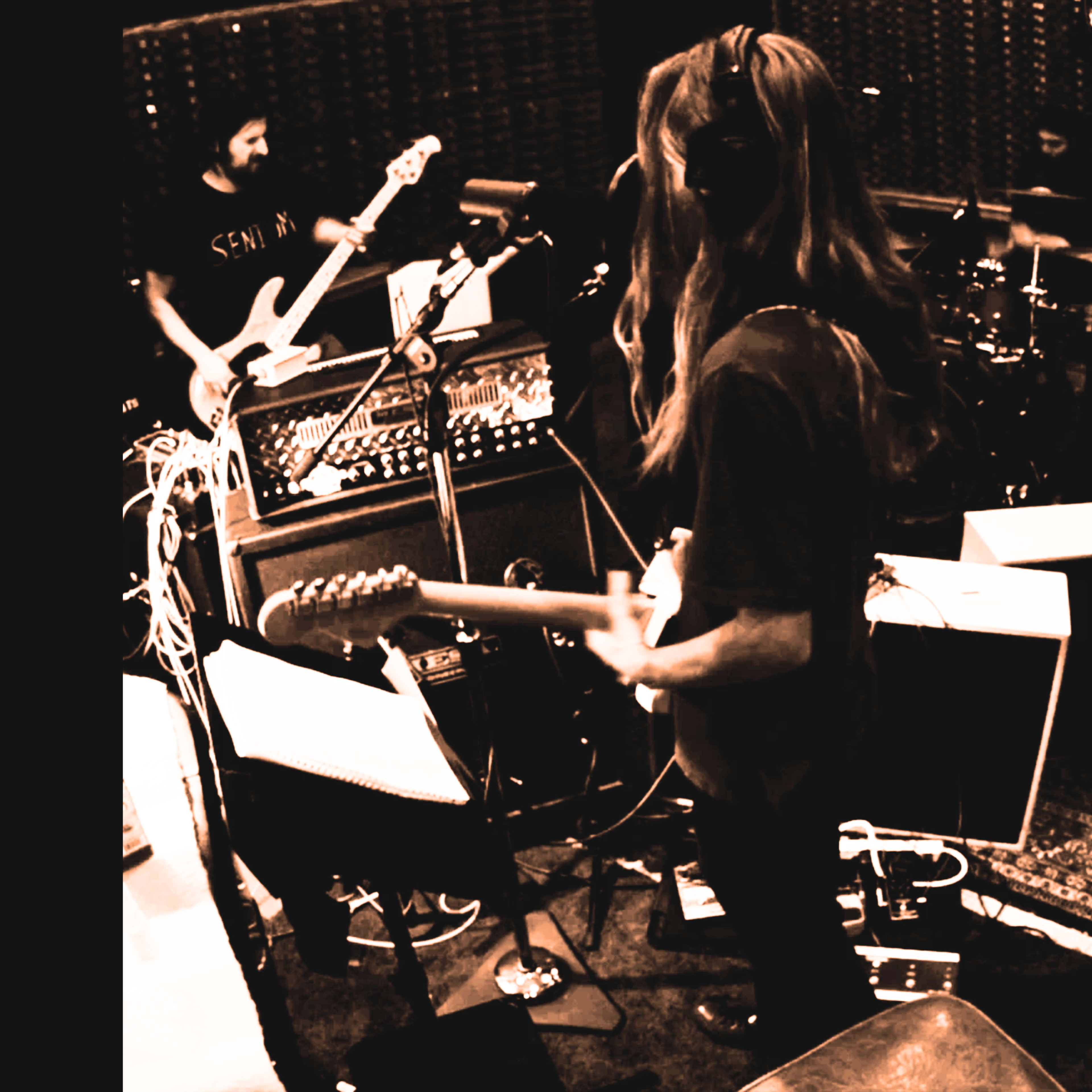 Musicians are recording in a studio with guitars, drums, and various equipment set up around them.