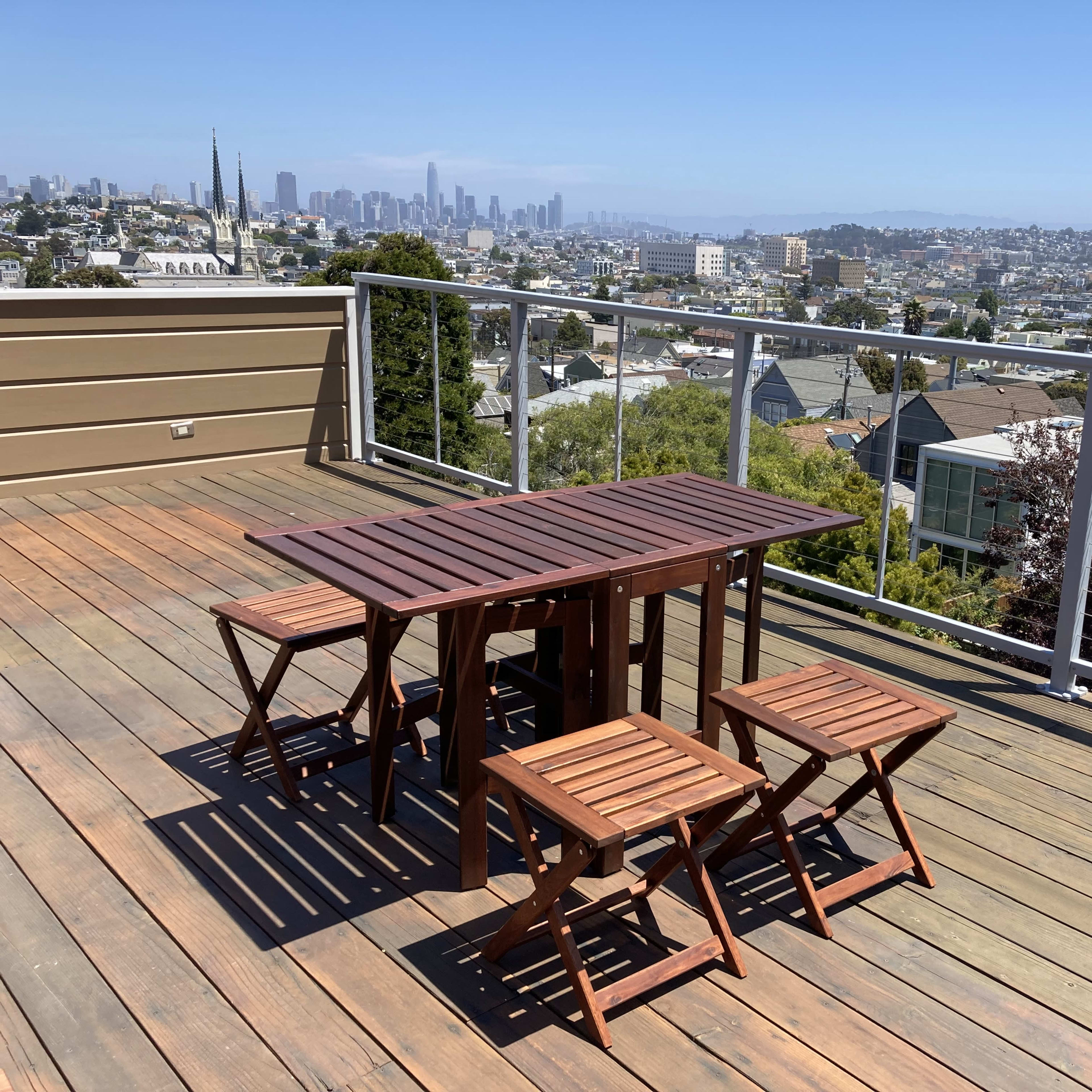 A wooden table with matching chairs is positioned on a deck overlooking a cityscape with distant buildings.