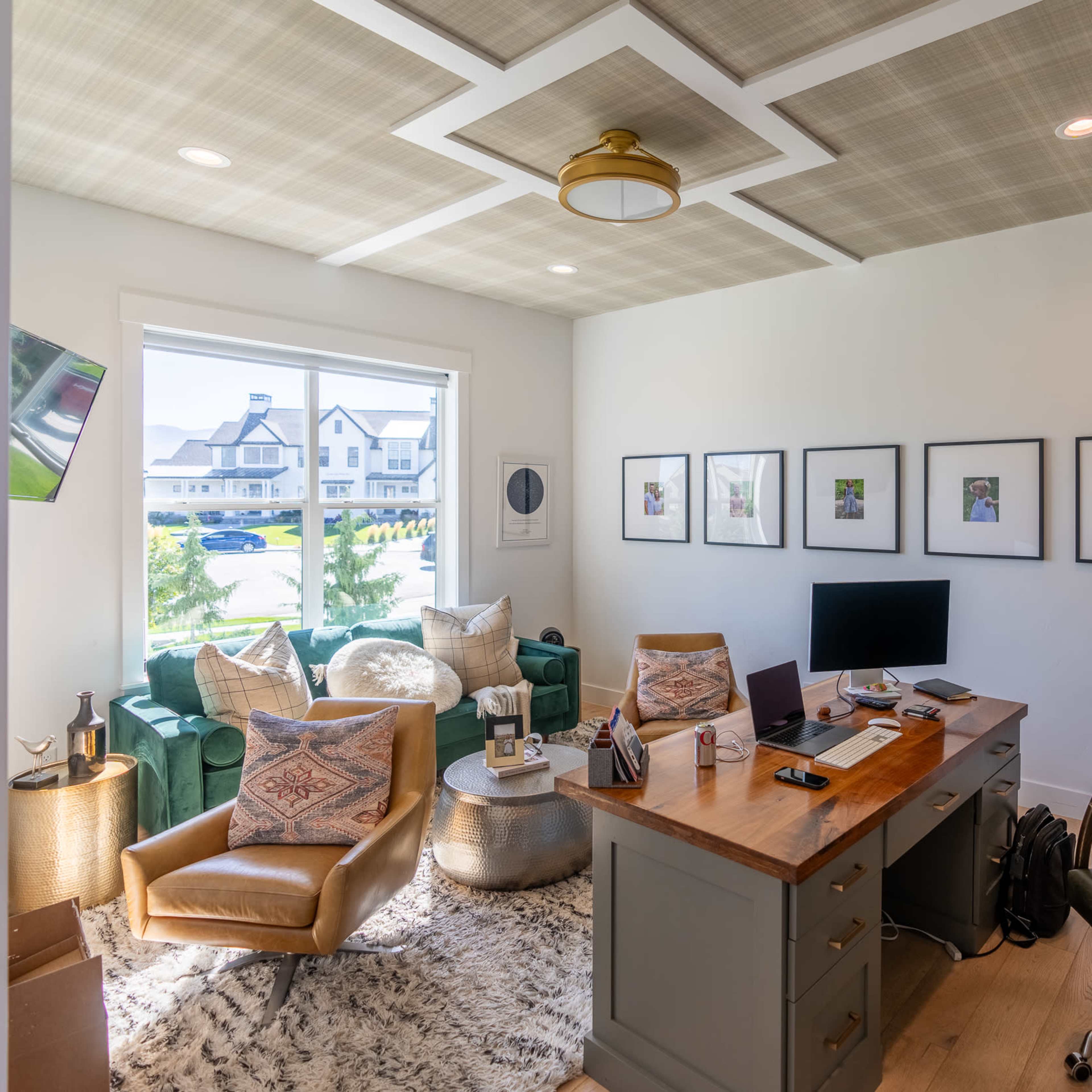 A modern home office featuring a wooden desk, several framed pictures on the wall, green armchairs, and a window letting in natural light.