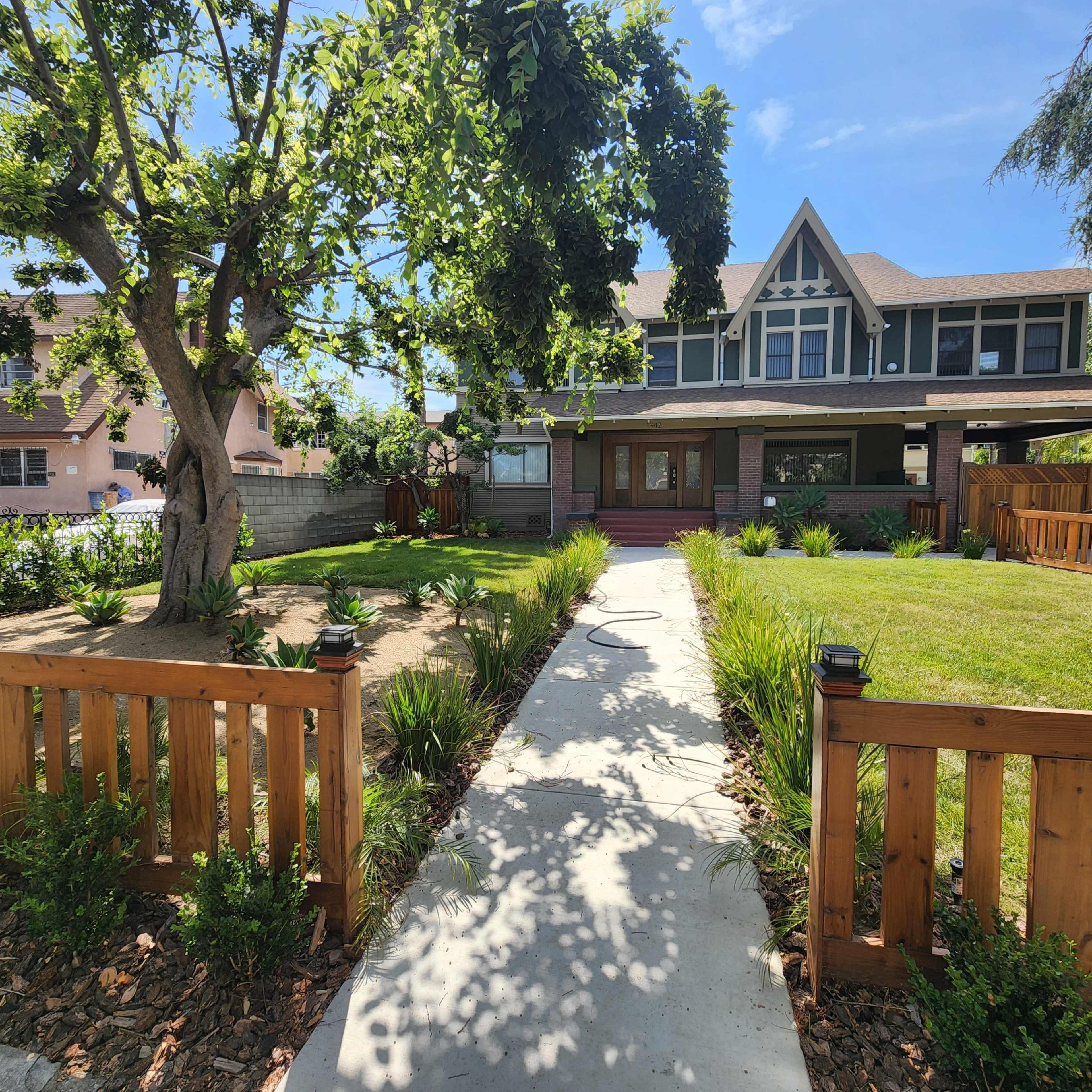 A concrete walkway leads through a landscaped yard to a large, multi-story house with a wraparound porch.