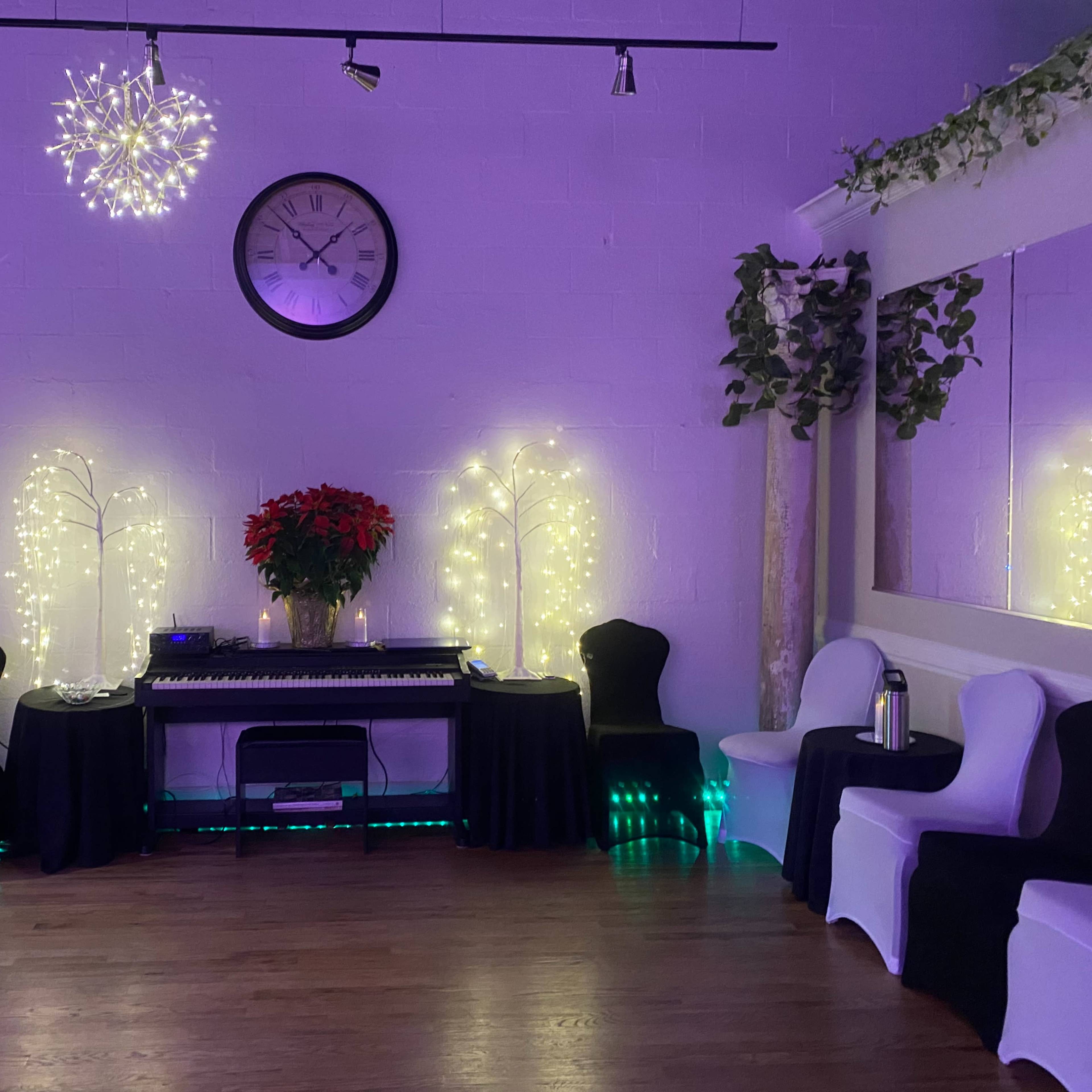A spacious dance studio with a wooden floor, red curtains, and decorative lighting, including chandeliers and a row of chairs along the walls.