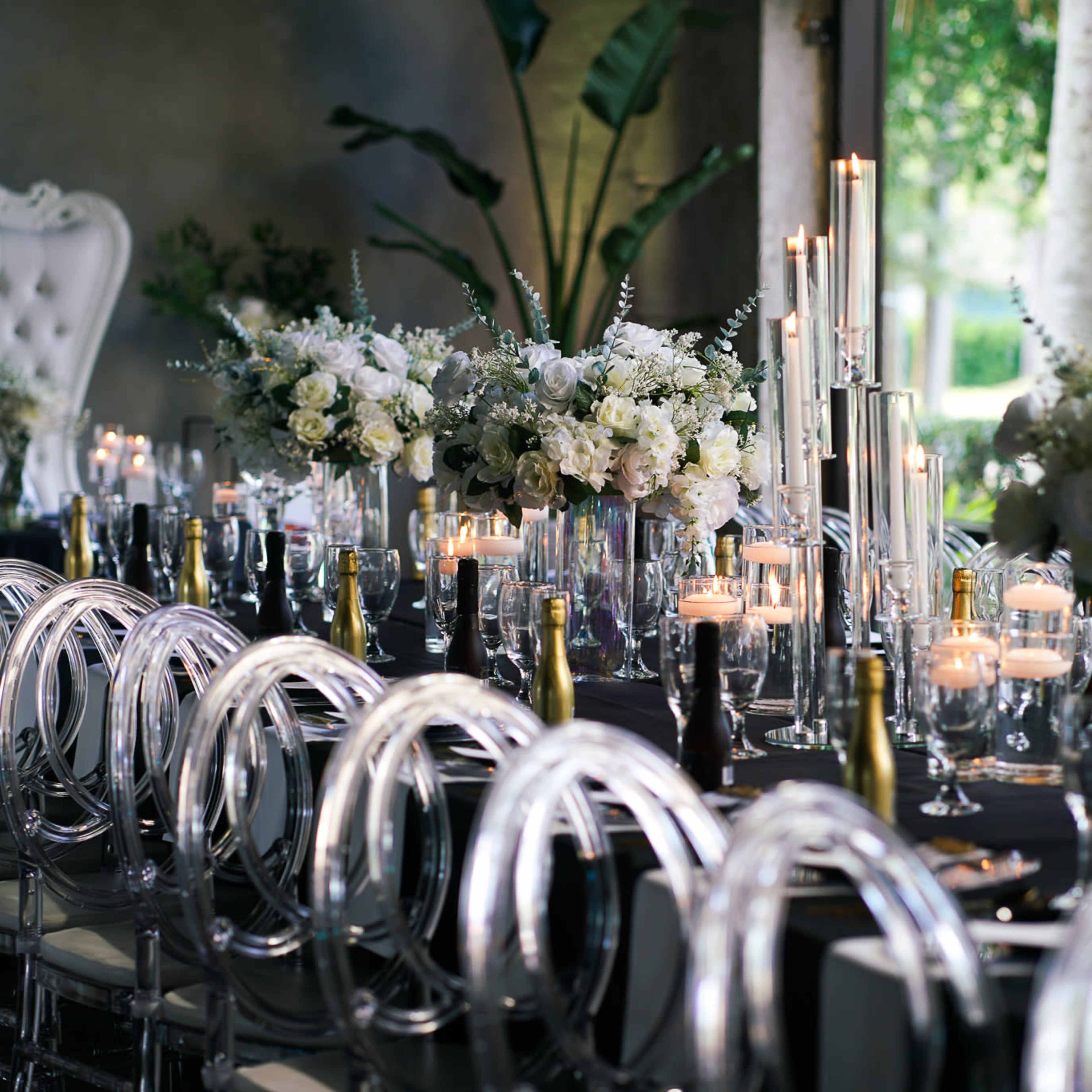 A long dining table is elegantly set with white flowers, candles, and clear glassware, surrounded by transparent chairs.