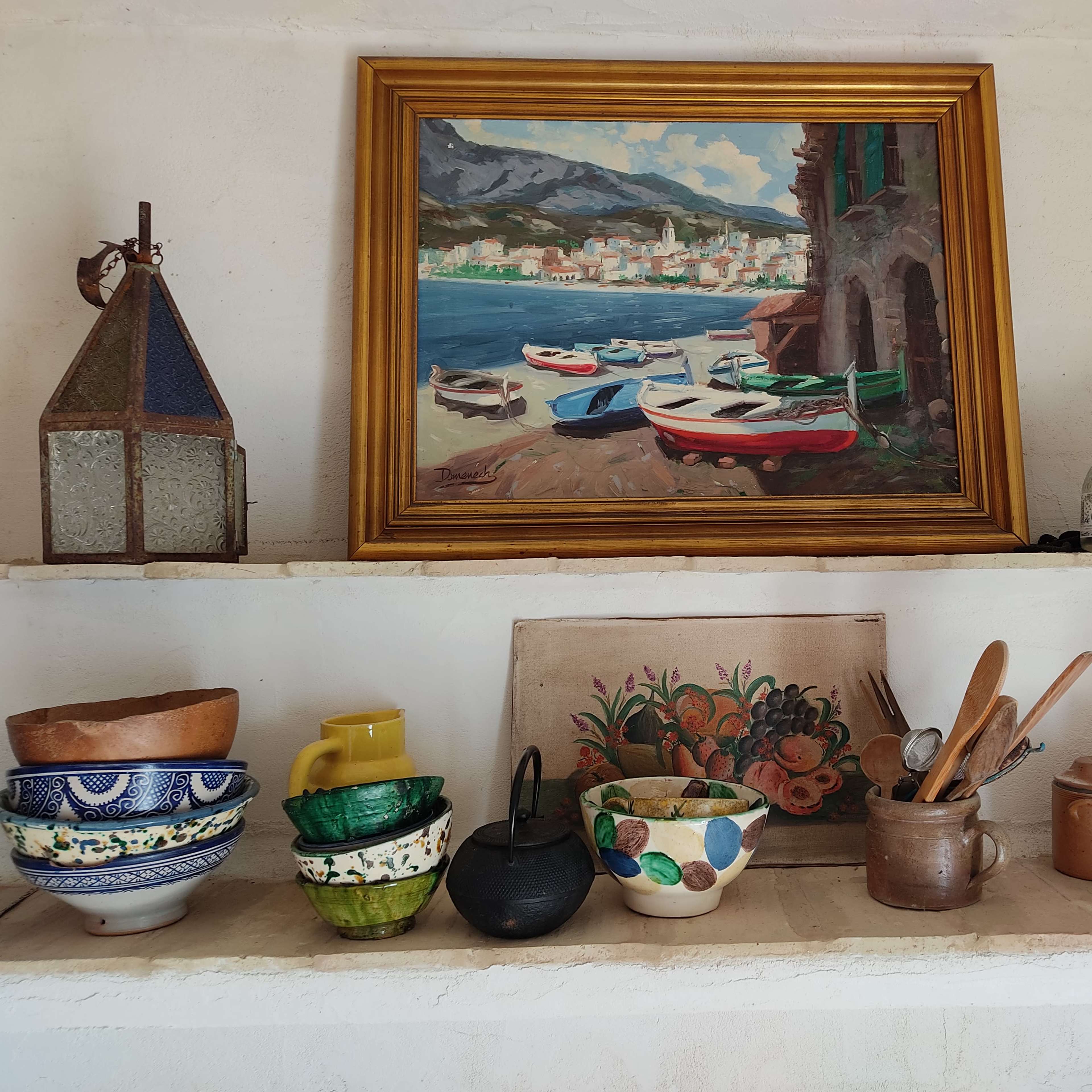 A collection of ceramic bowls, jars, and kitchen utensils are arranged on shelves beneath a landscape painting of boats by a coastal town.