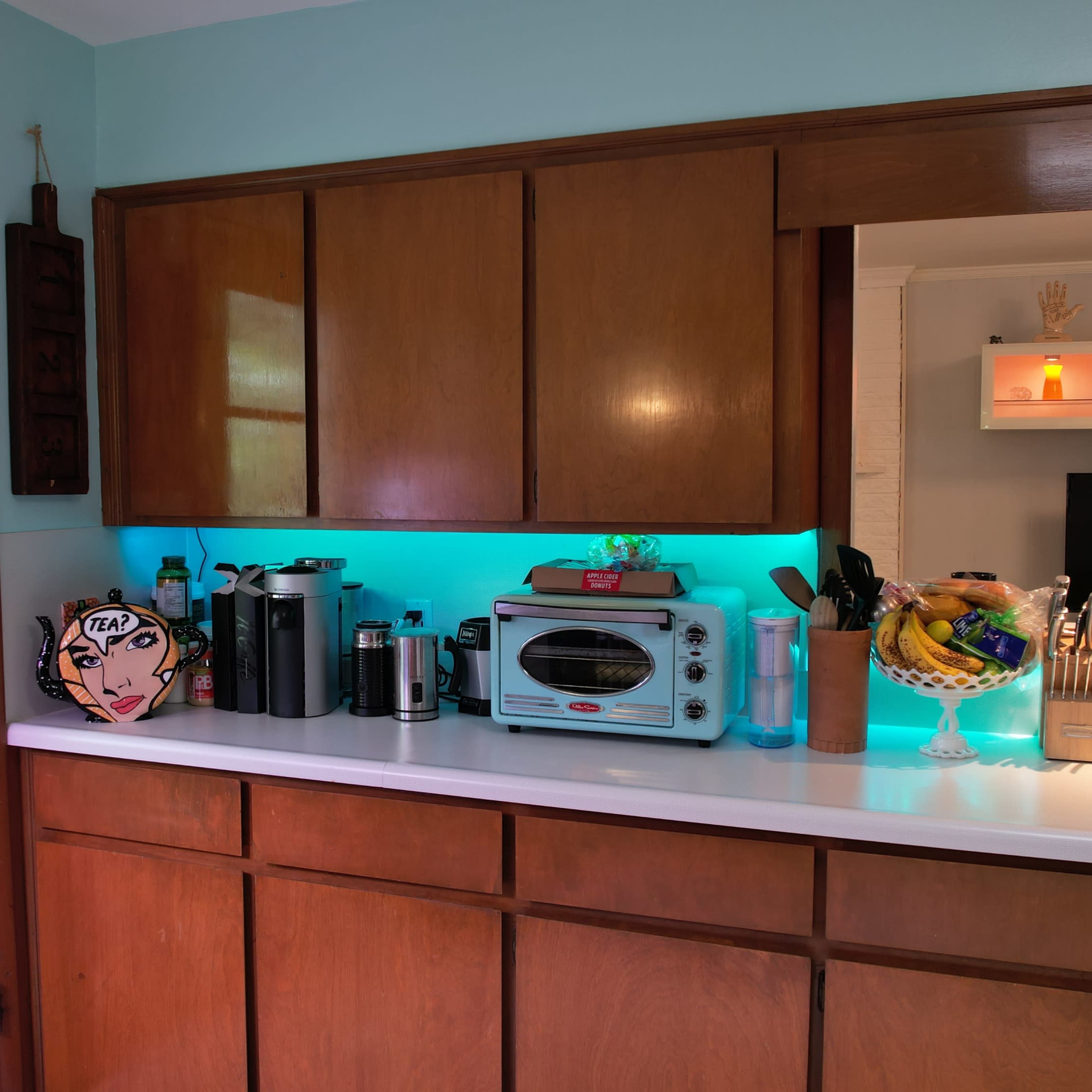 A kitchen with wooden cabinets, a countertop featuring various appliances, and blue LED lighting along the backsplash.