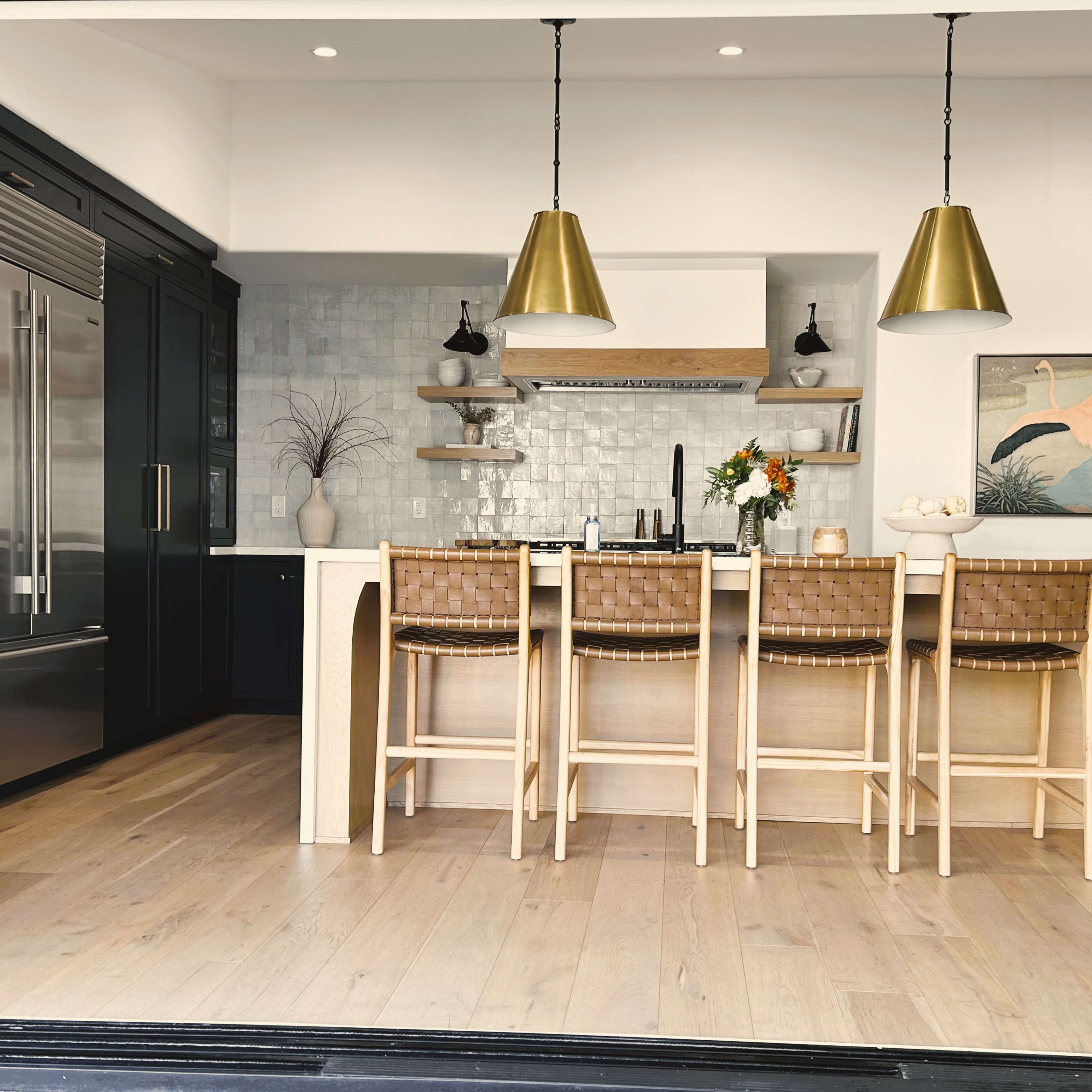 A modern kitchen featuring a light wood island with four bar stools, stainless steel appliances, and pendant lights above.