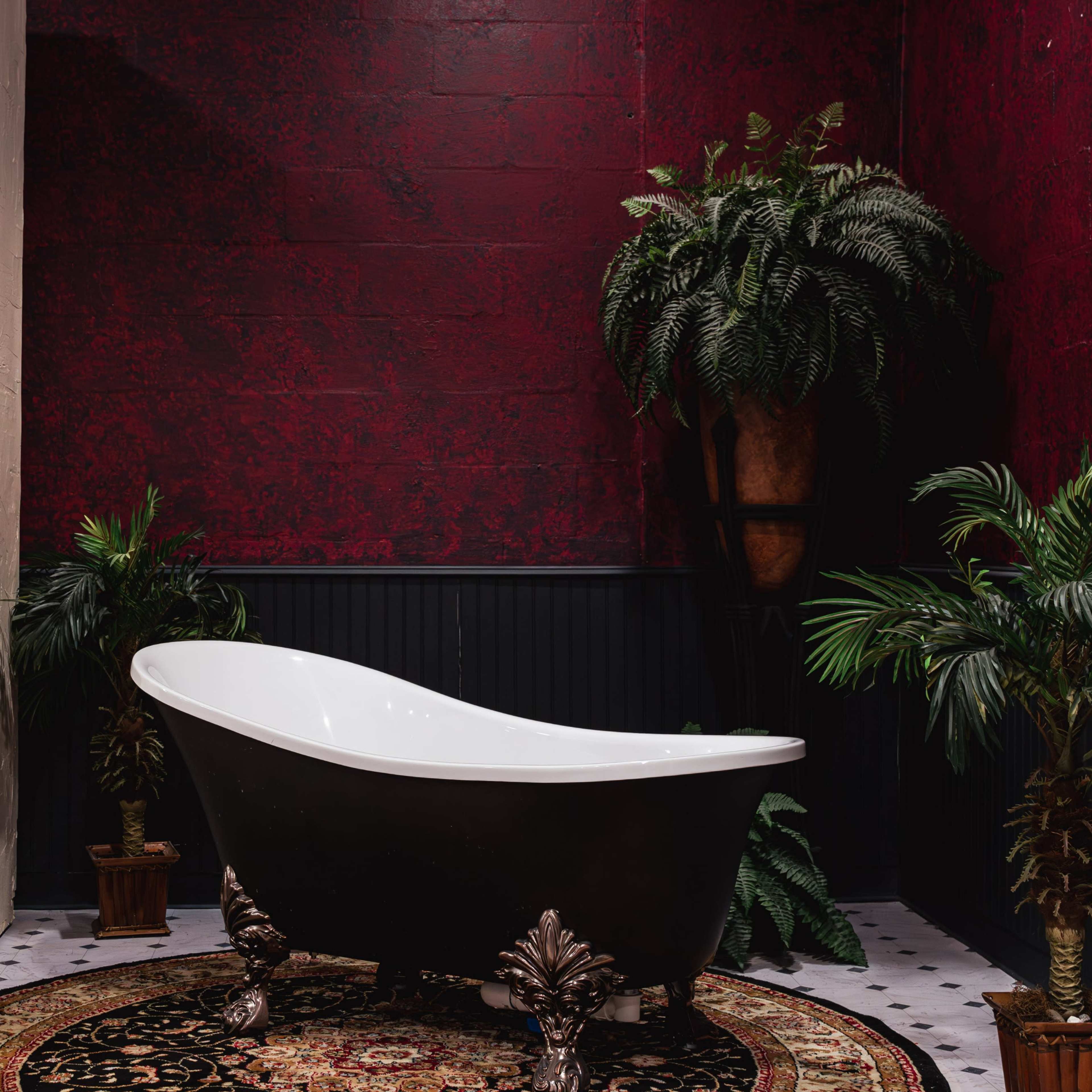 A freestanding black bathtub is situated on a circular patterned rug in a room with deep red walls and lush green plants.