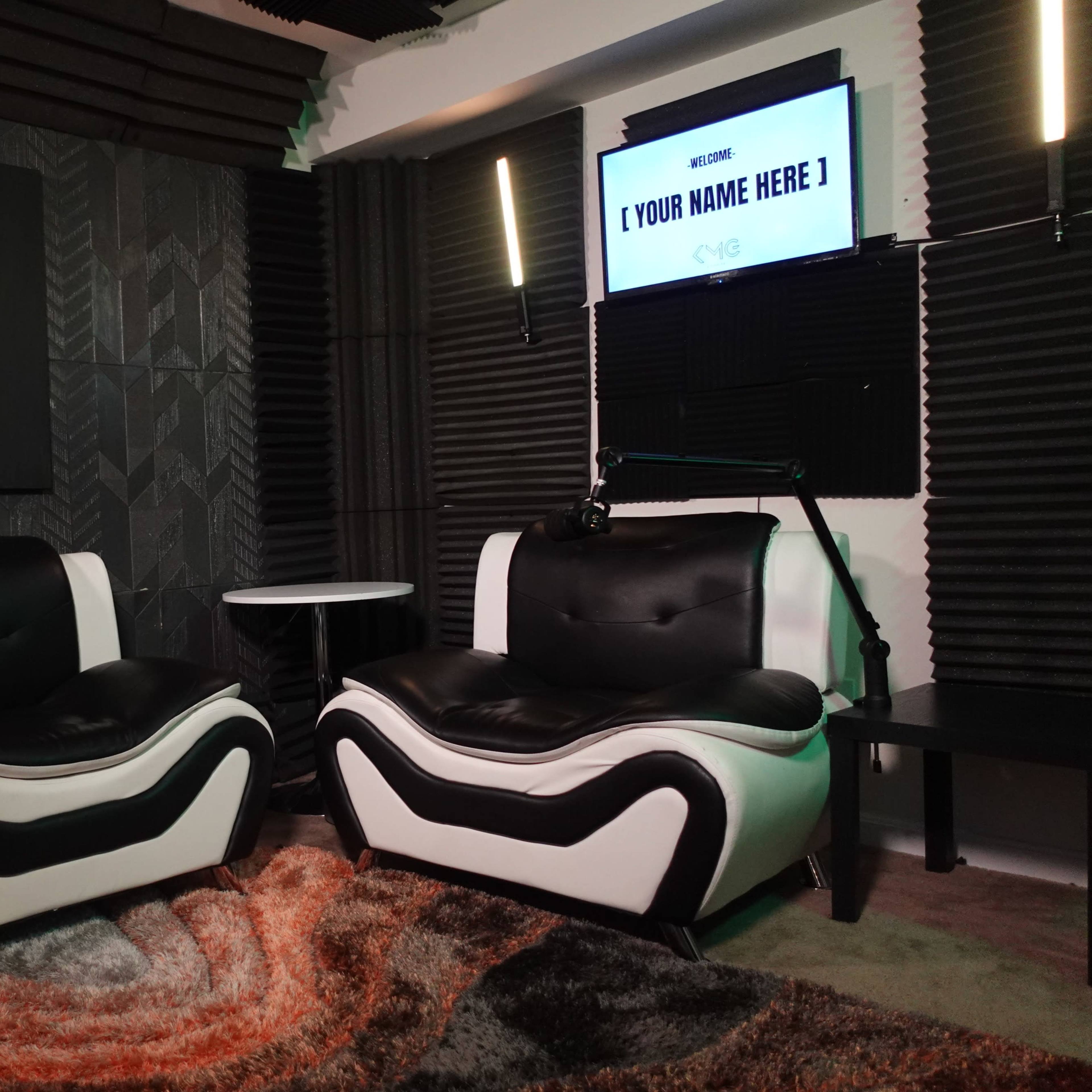 A modern podcast studio featuring two black and white armchairs, a wall-mounted screen with a customizable title, and acoustic paneling behind the chairs.