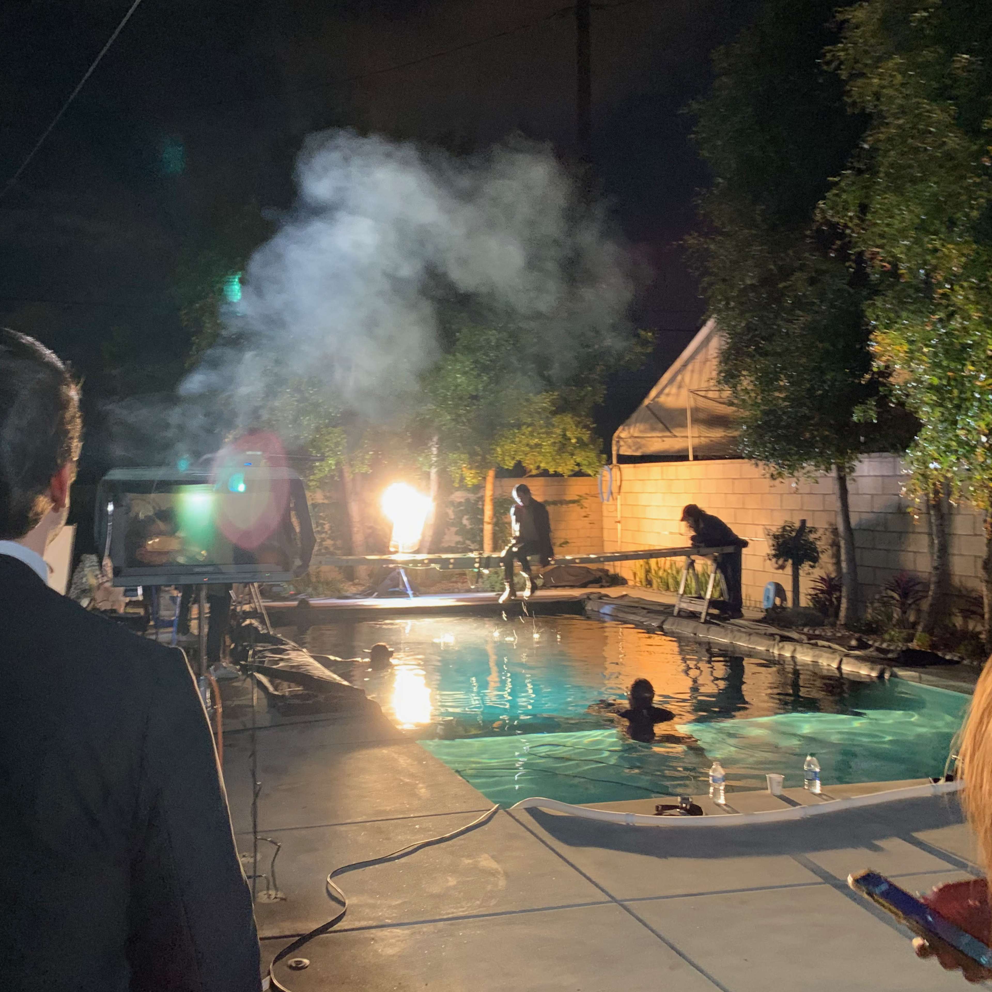 A person in formal attire stands by a pool as smoke billows from a device near the water while others are visible in the background.