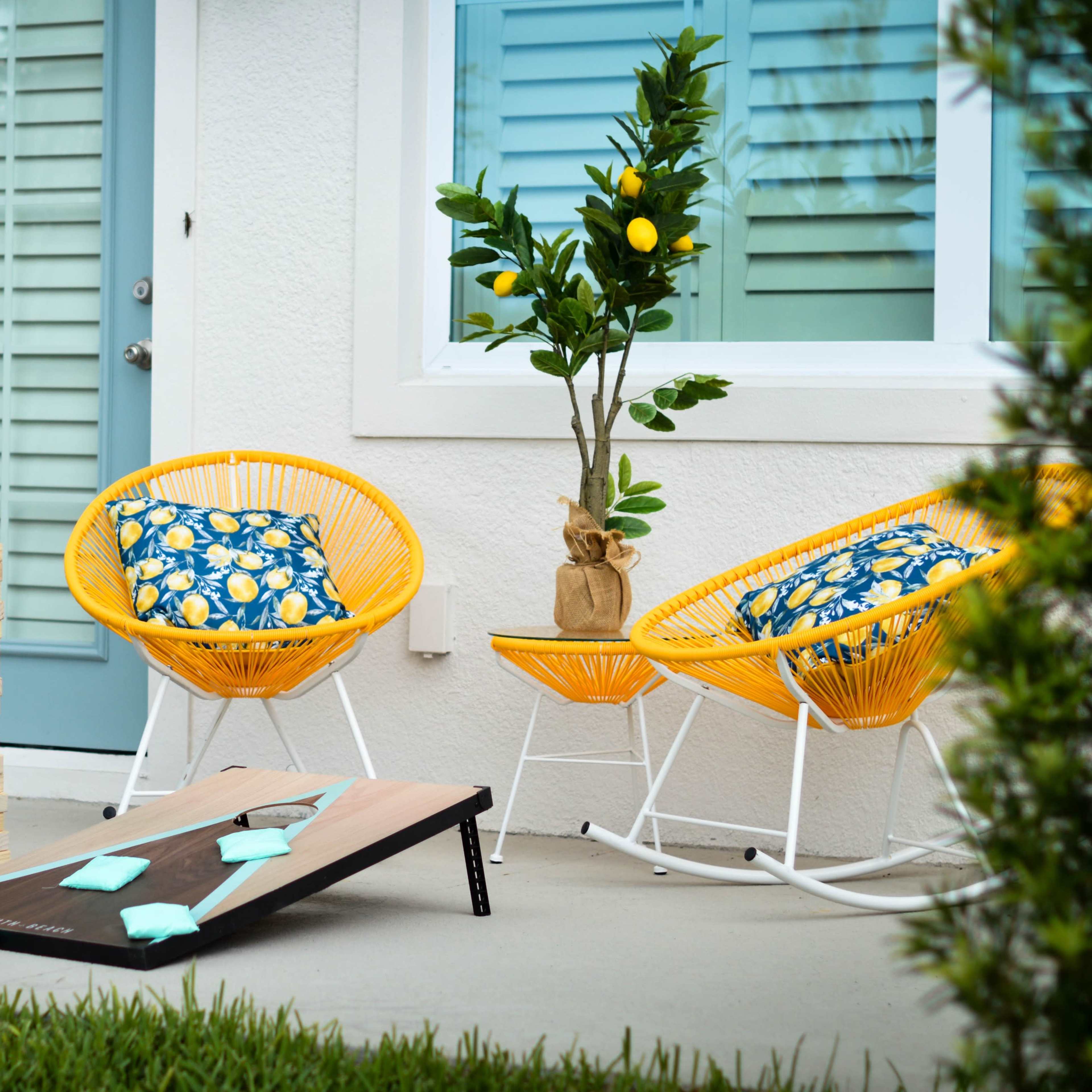 The scene features two bright yellow chairs with floral cushions, a small potted tree, and a stack of wooden blocks beside a cornhole game on a patio.