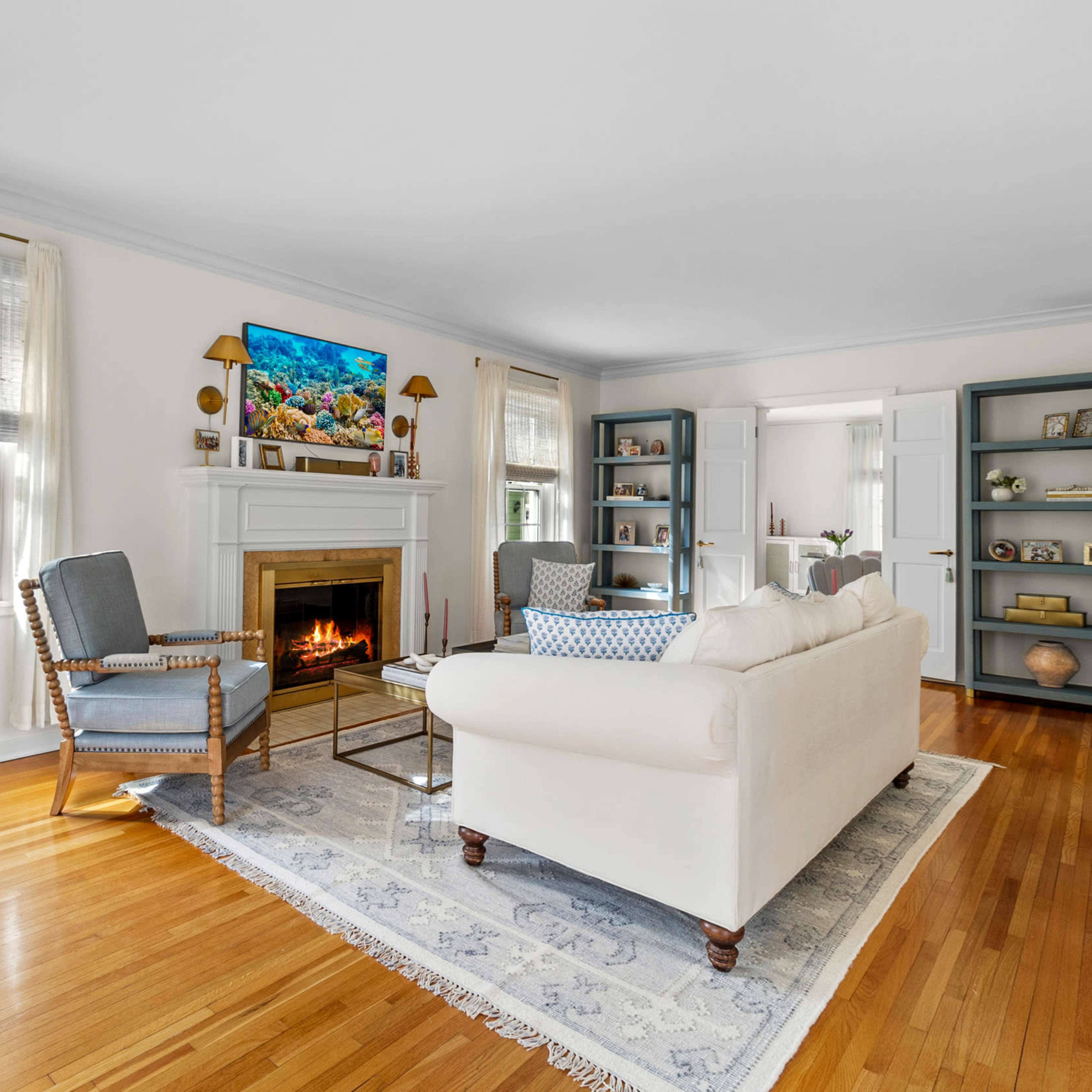 A bright living room features a white sofa facing a fireplace, flanked by two chairs, with shelves and a television on the walls.