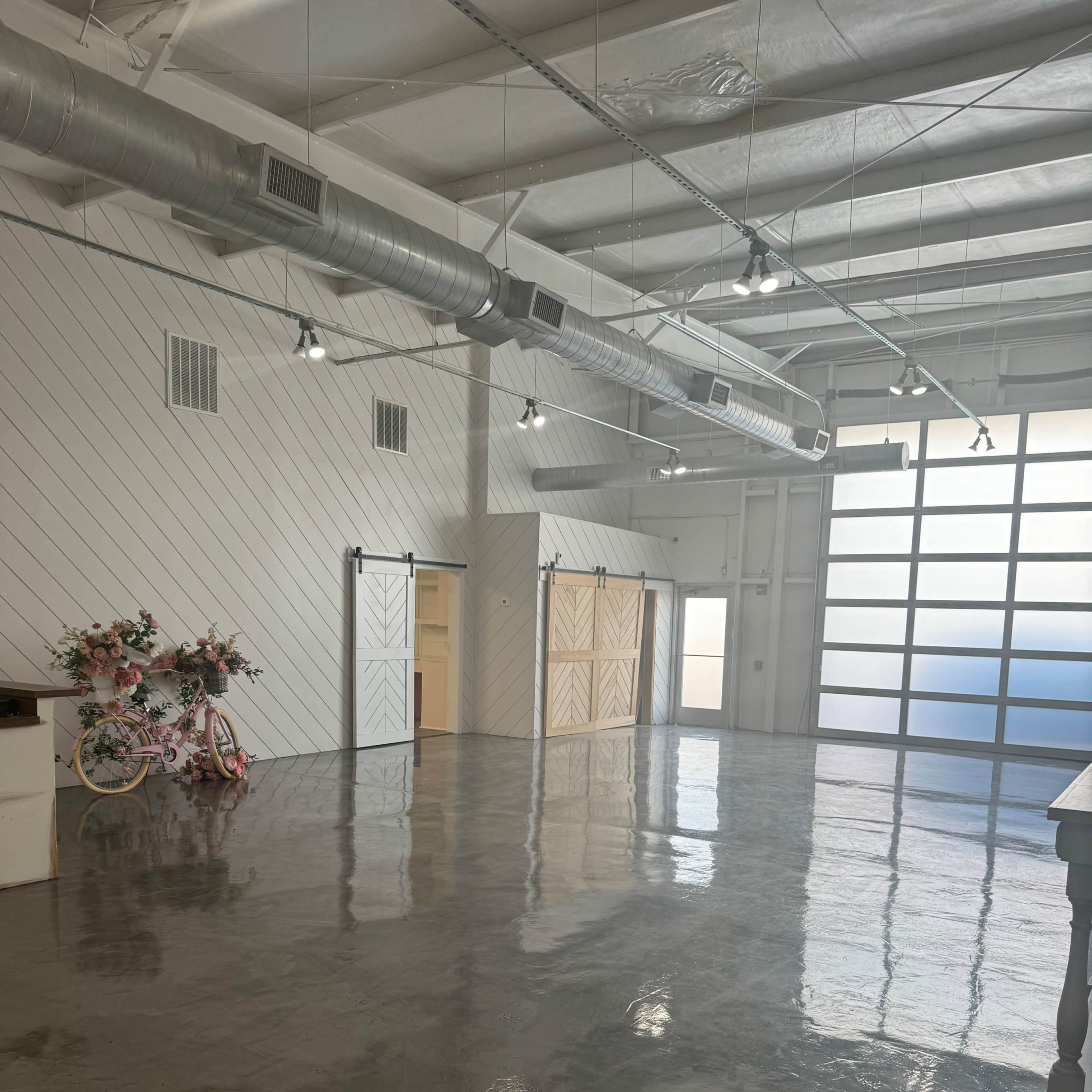 A wide, empty room with polished concrete floors, white walls, and large windows, featuring a pink bicycle and floral decorations in one corner.