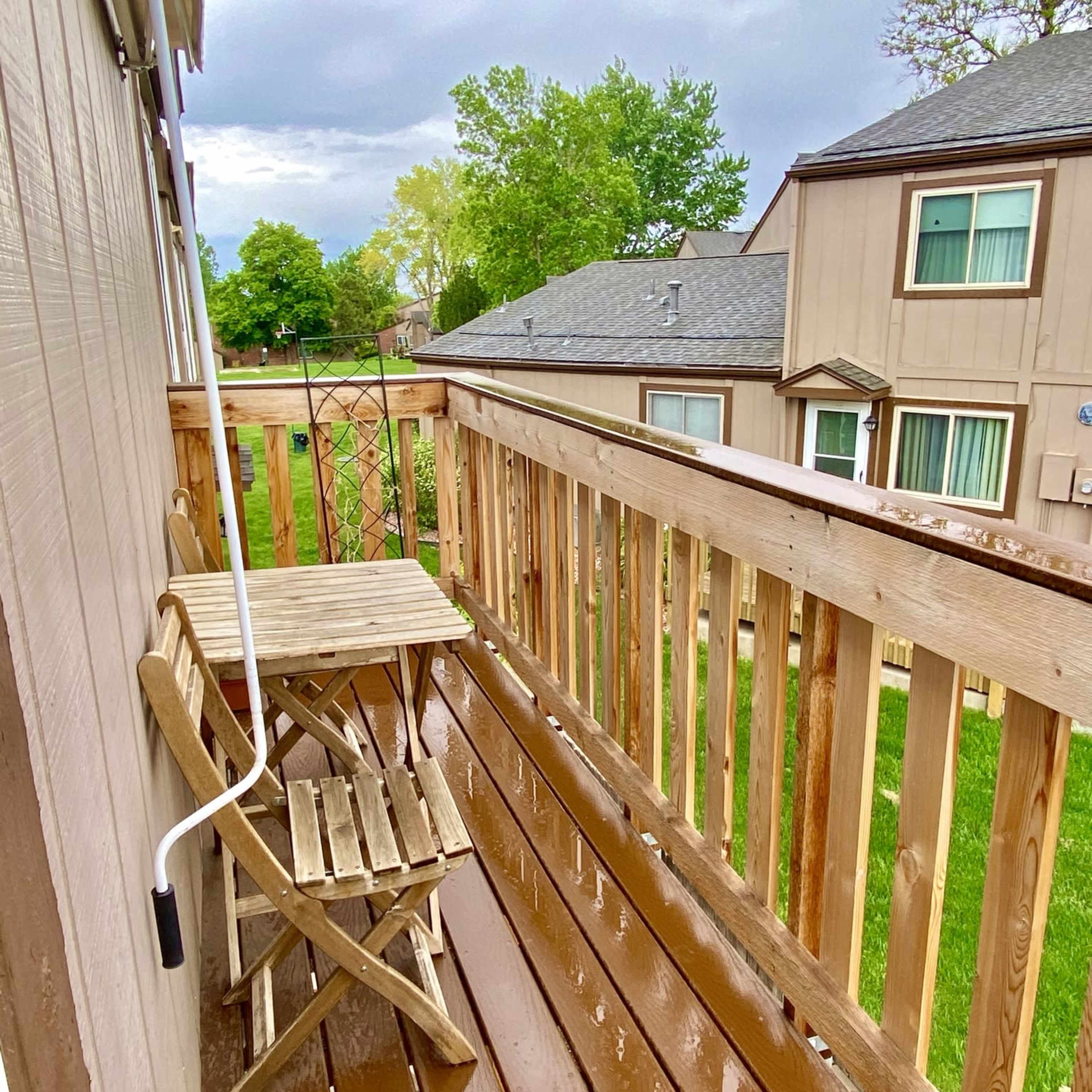 A wooden balcony features a small table and chairs overlooking a green yard and nearby houses under a cloudy sky.