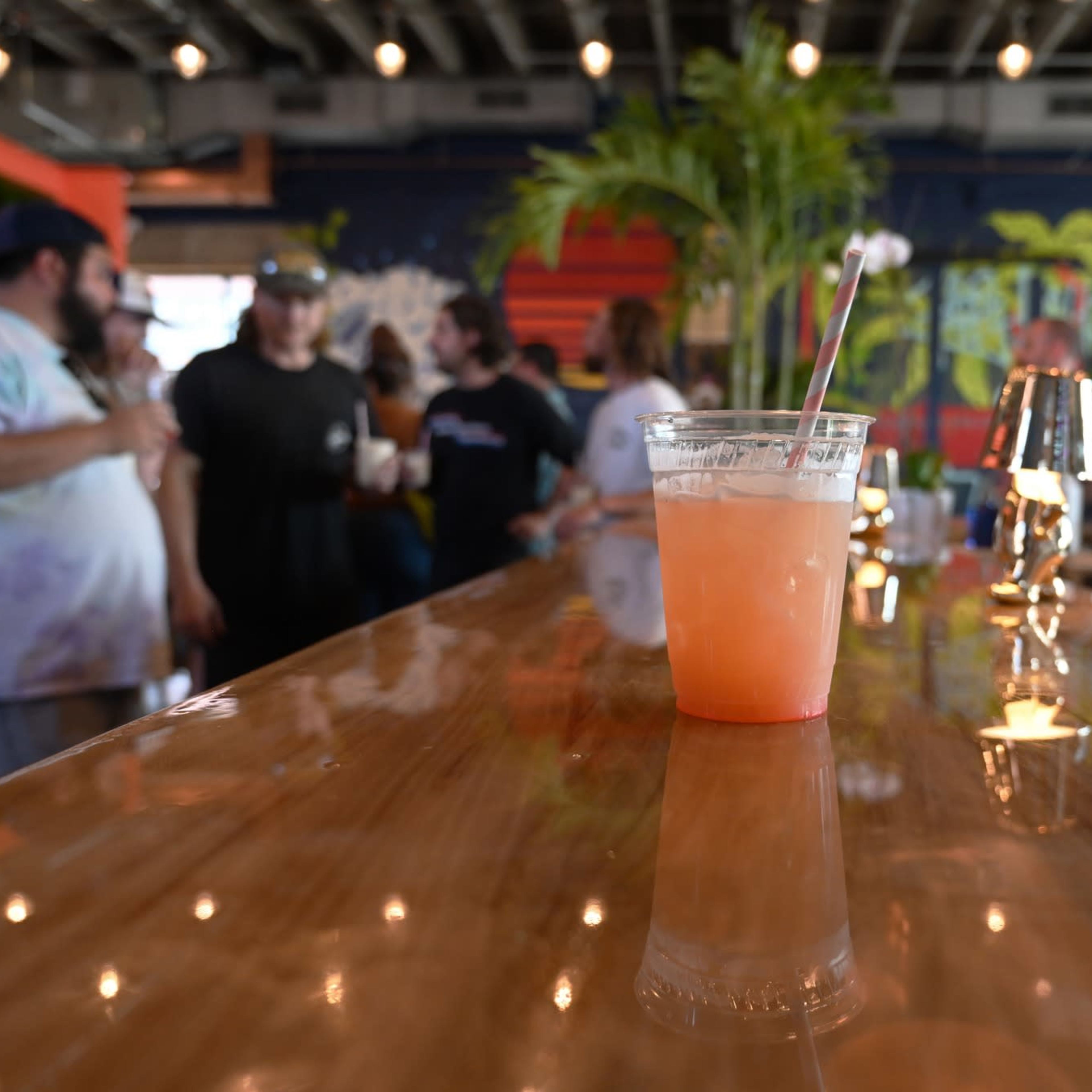 A clear plastic cup filled with a pink drink sits on a polished wooden bar, with a group of people conversing in the background.