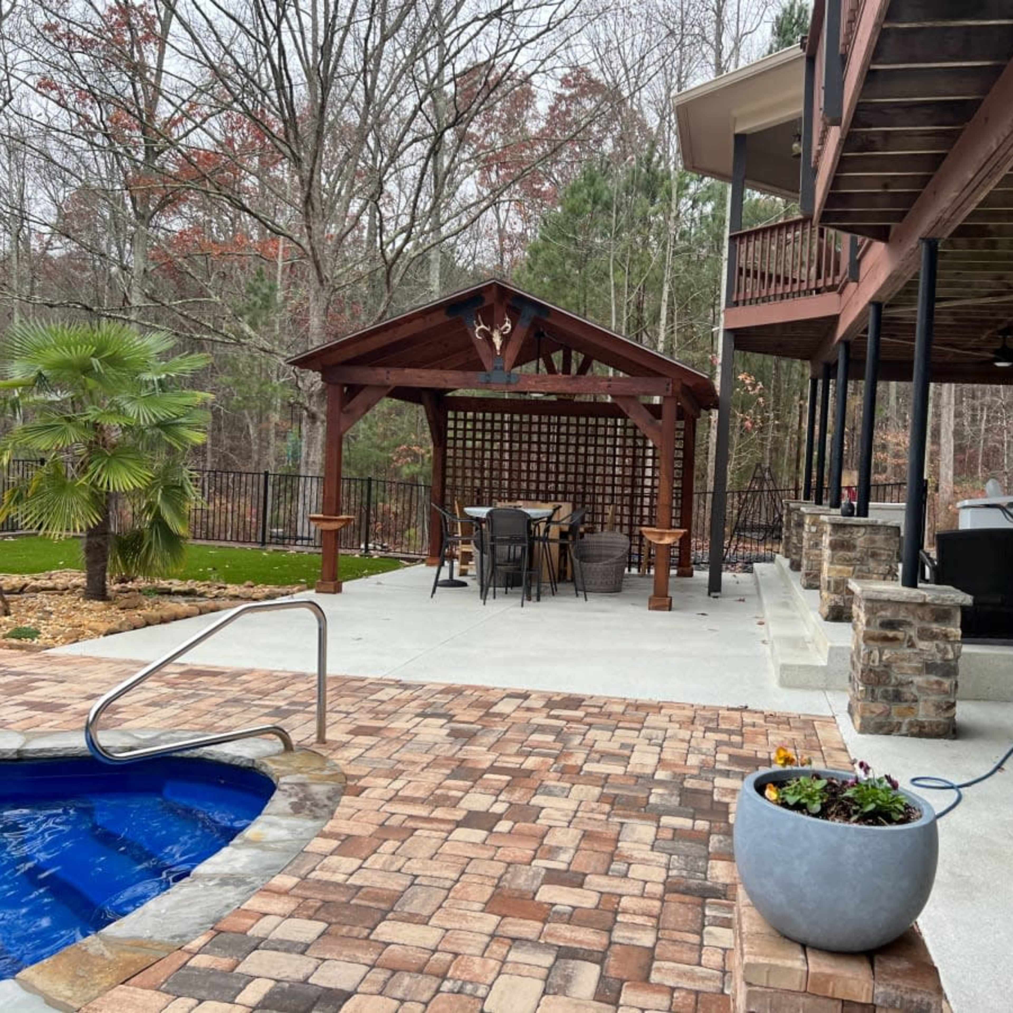 A patio area features a swimming pool, a seating arrangement under a wooden gazebo, and stone planters alongside the house.