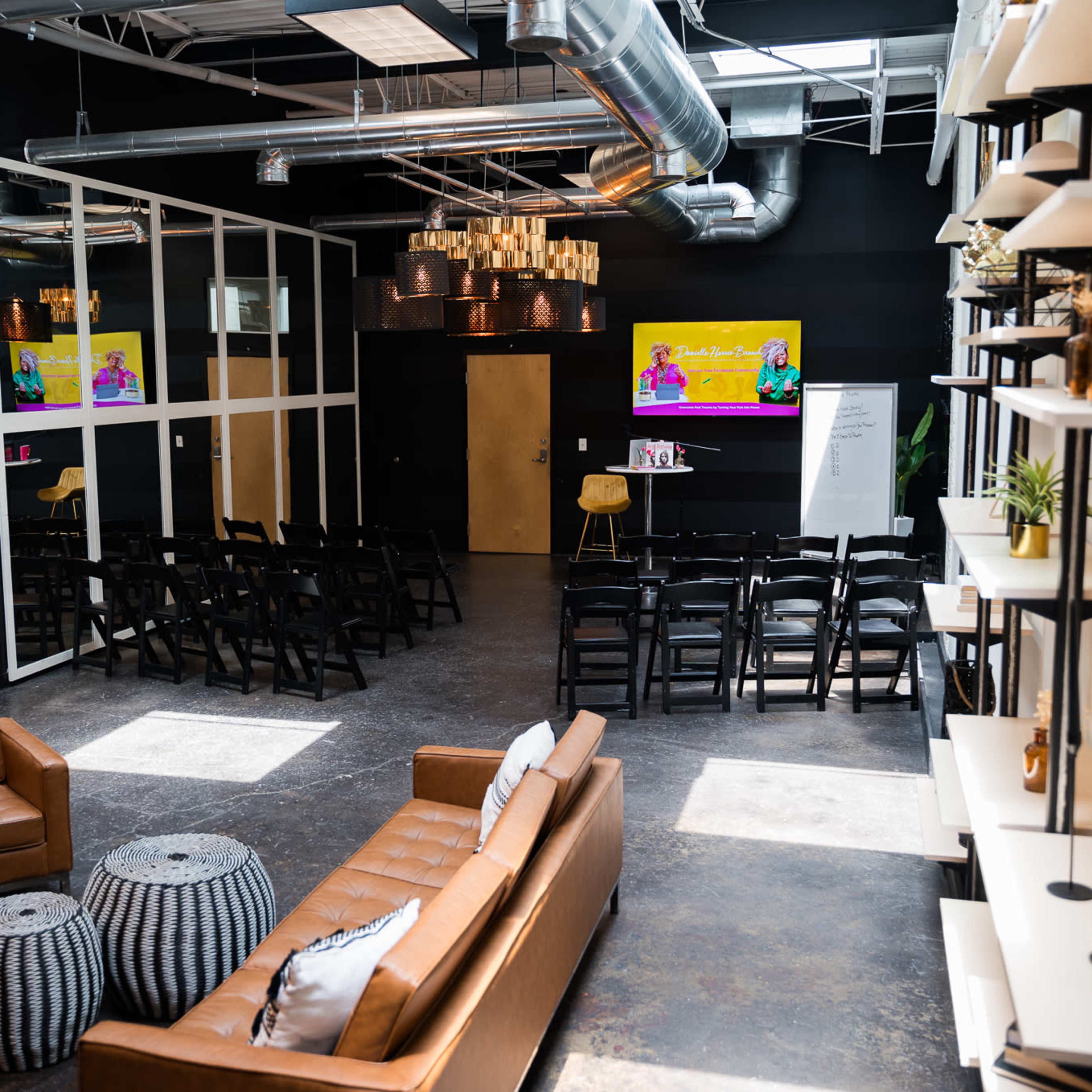 A modern, open event space featuring rows of chairs, a brown leather sofa, and two large screens displaying colorful presentations.