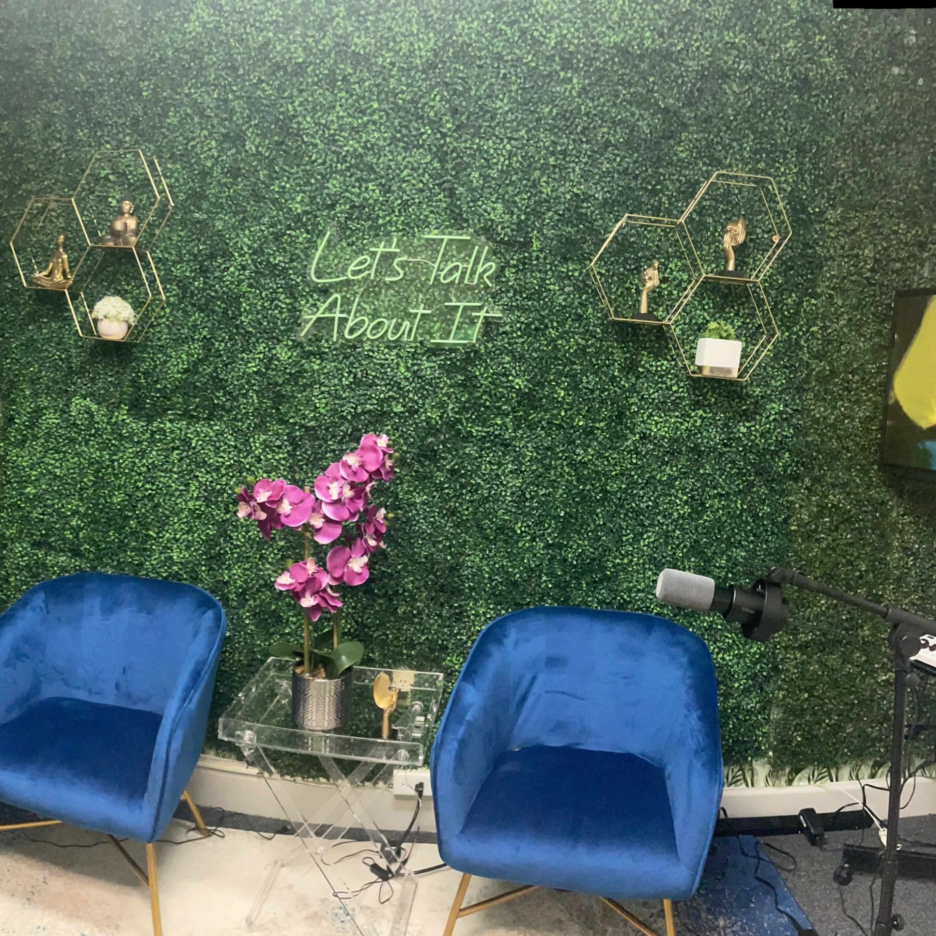 A modern interview or discussion space featuring two blue velvet chairs, two barstools, a green plant wall, and decorative wall art.