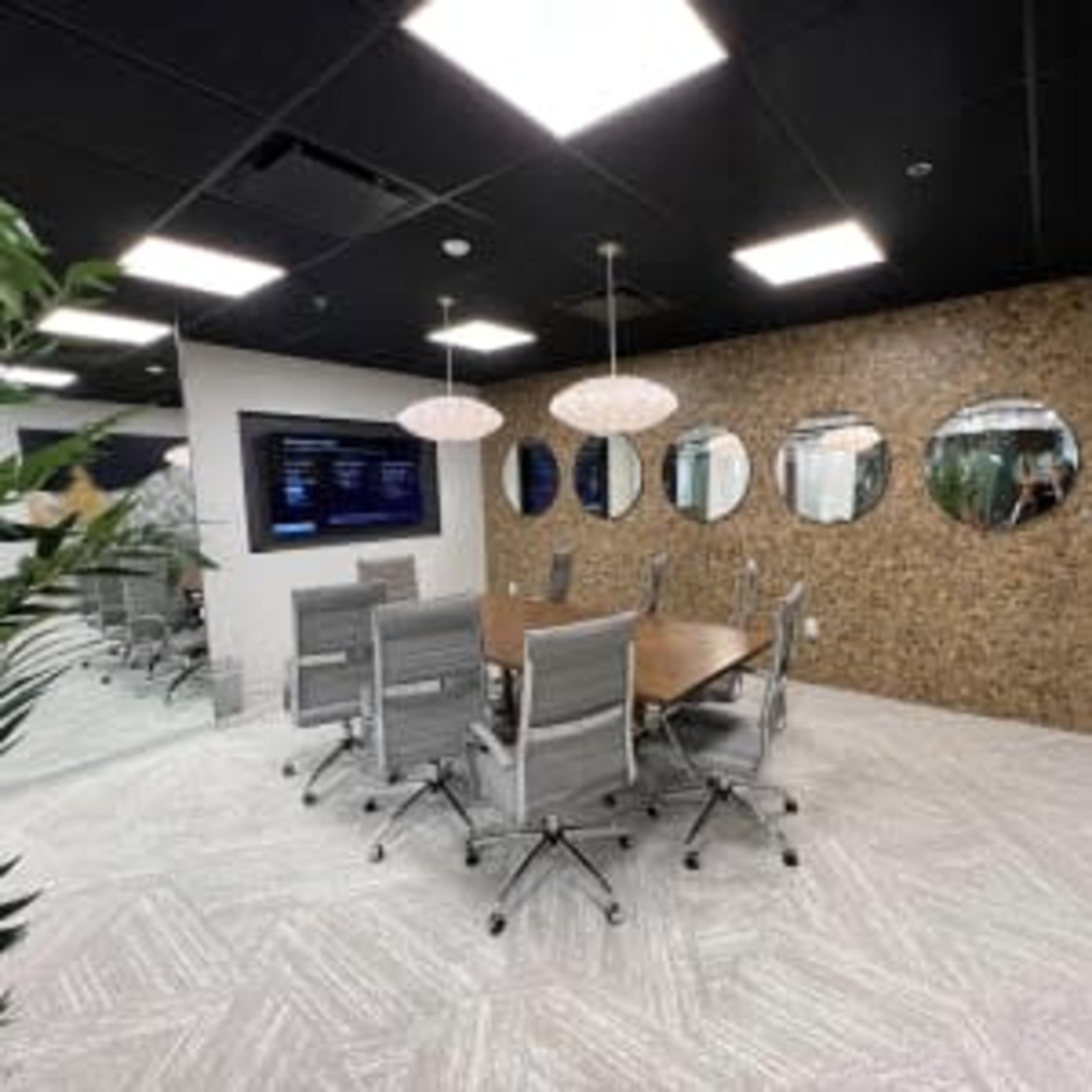 A modern conference room features a large table surrounded by gray chairs, with a wall of decorative mirrors and a display screen.