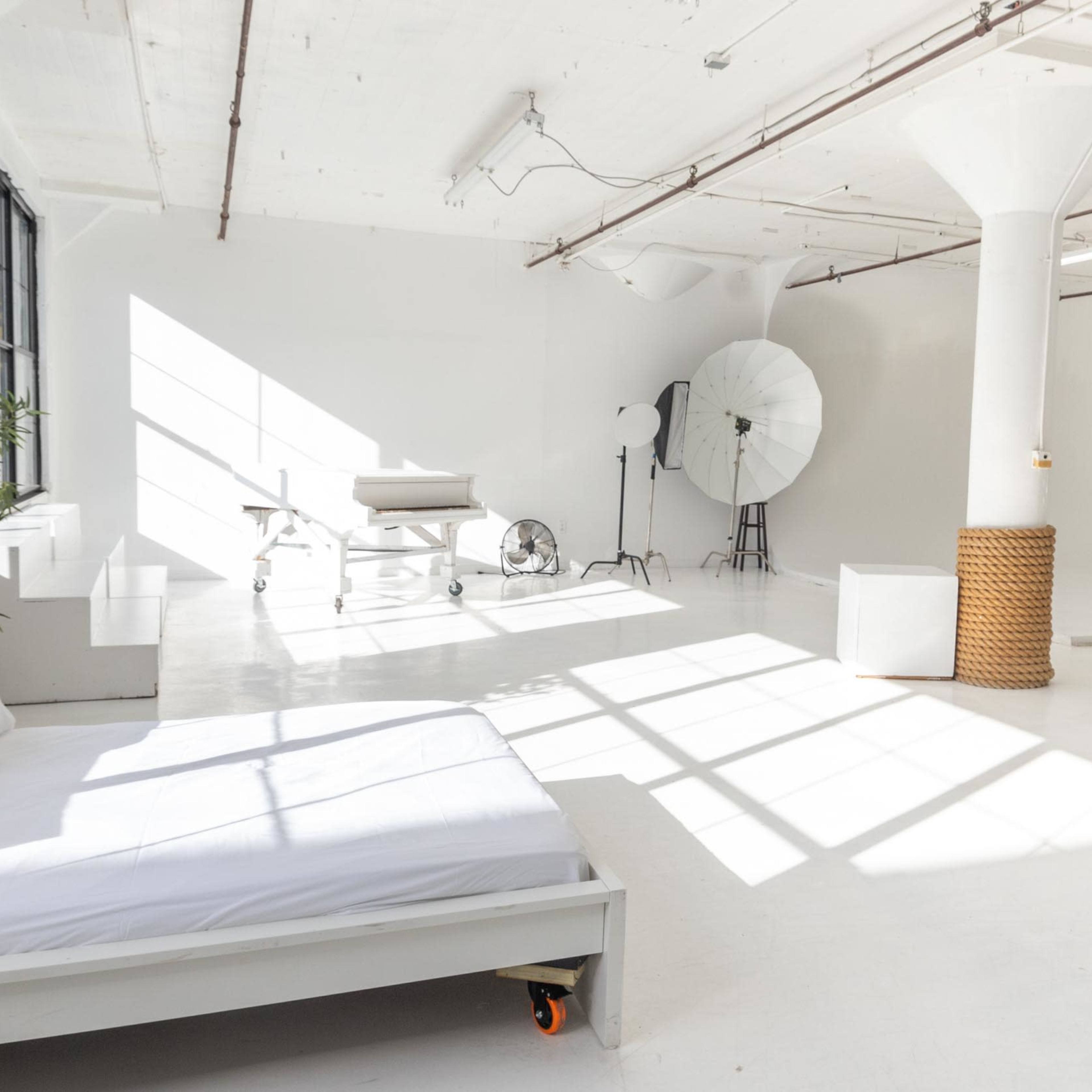A bright, minimalistic studio space featuring a bed, a photography setup with softboxes, and large windows casting shadows on the white floor.