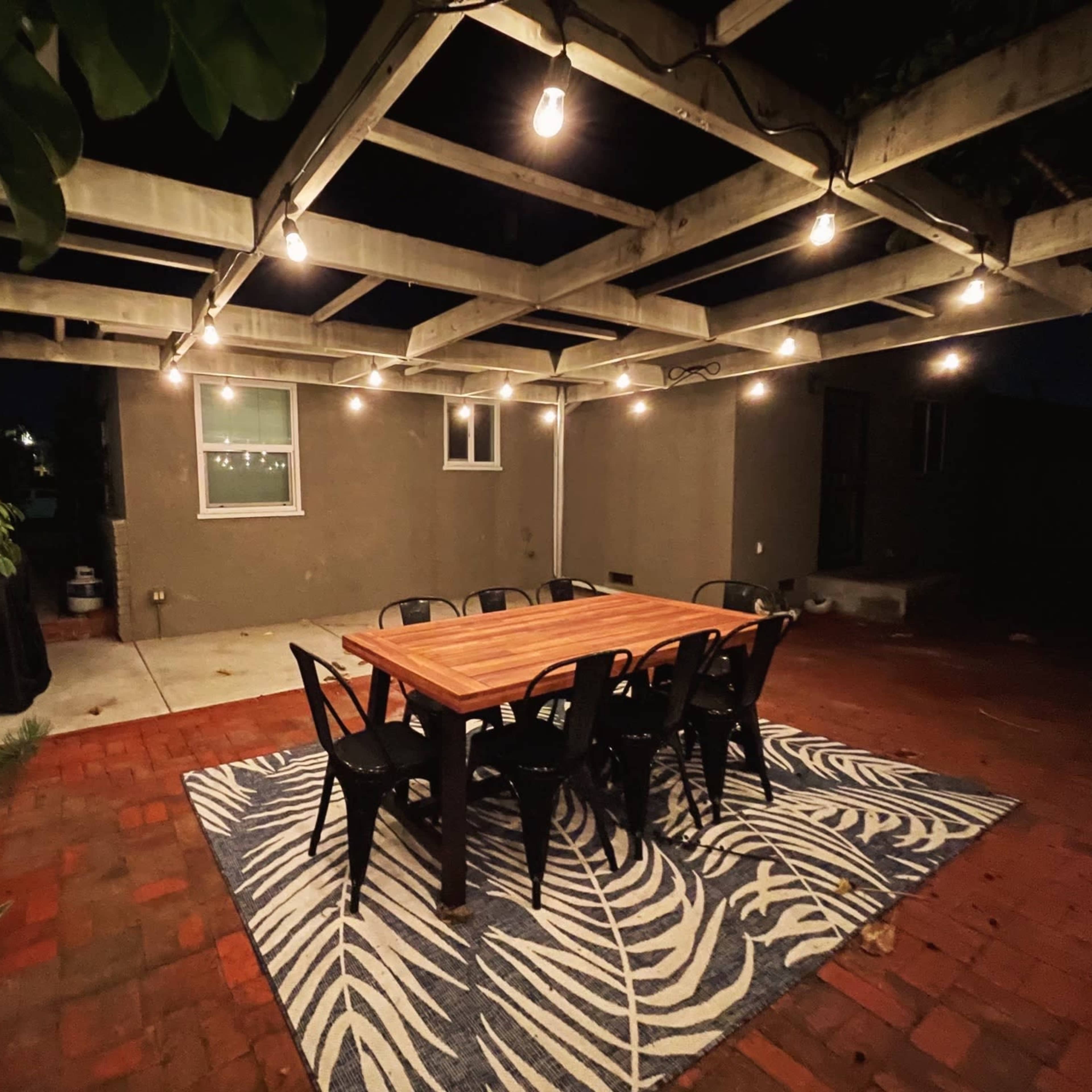 A wooden dining table surrounded by chairs is set under a pergola adorned with string lights in a backyard patio.