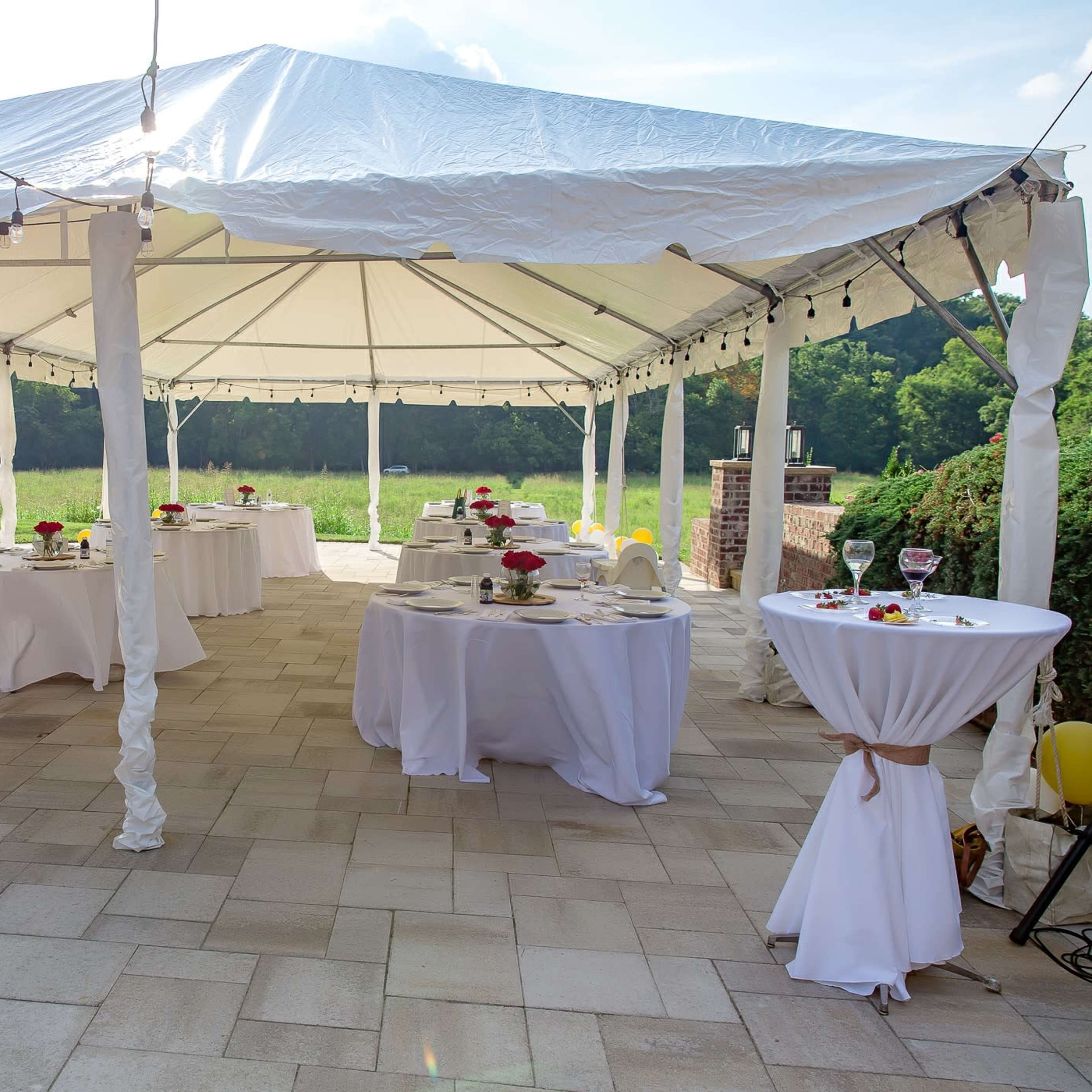 A large white tent with several round tables set for a gathering sits on a stone patio overlooking a green field.