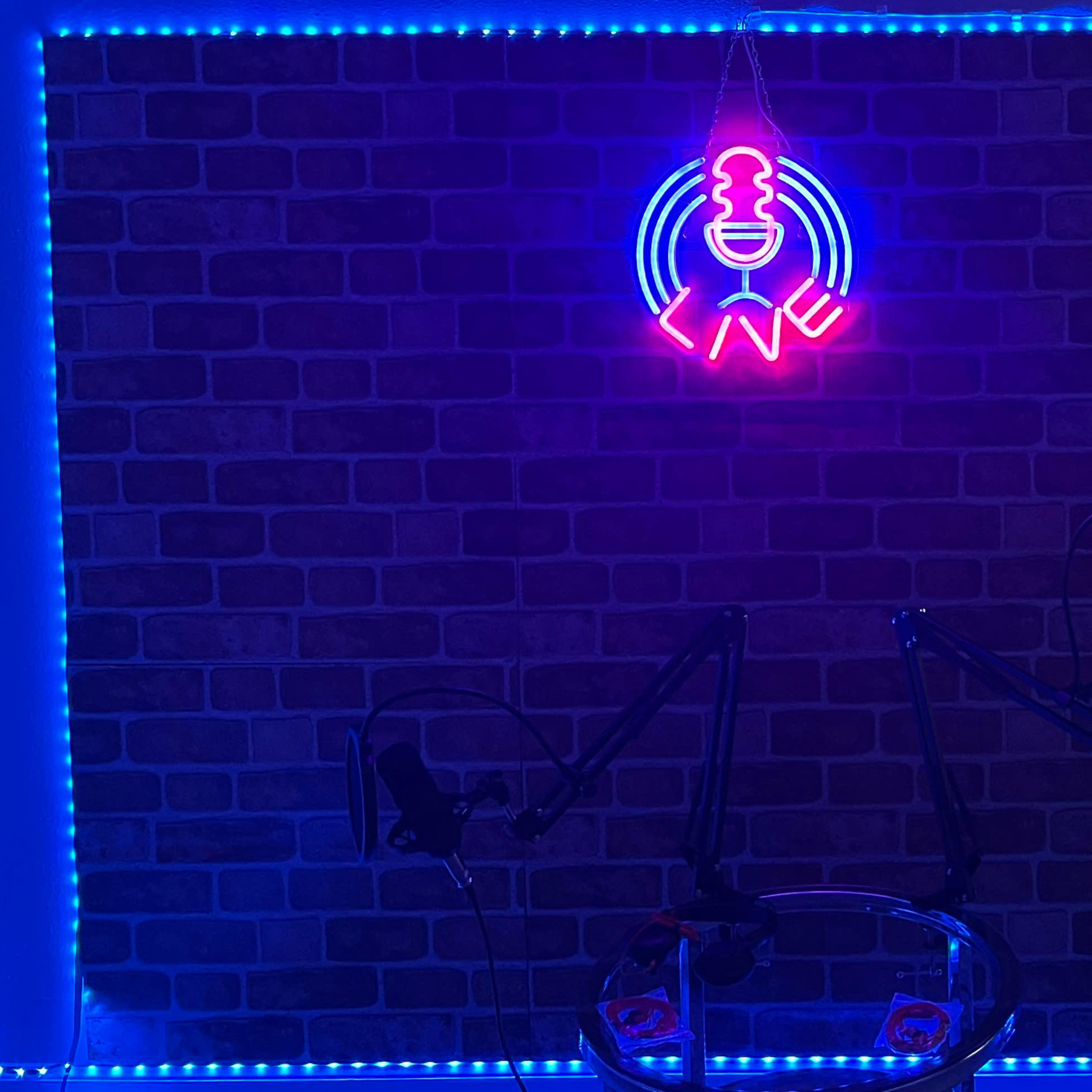 A podcast studio with a neon microphone sign and blue LED lights illuminating a brick wall, along with two microphone stands and a glass table.