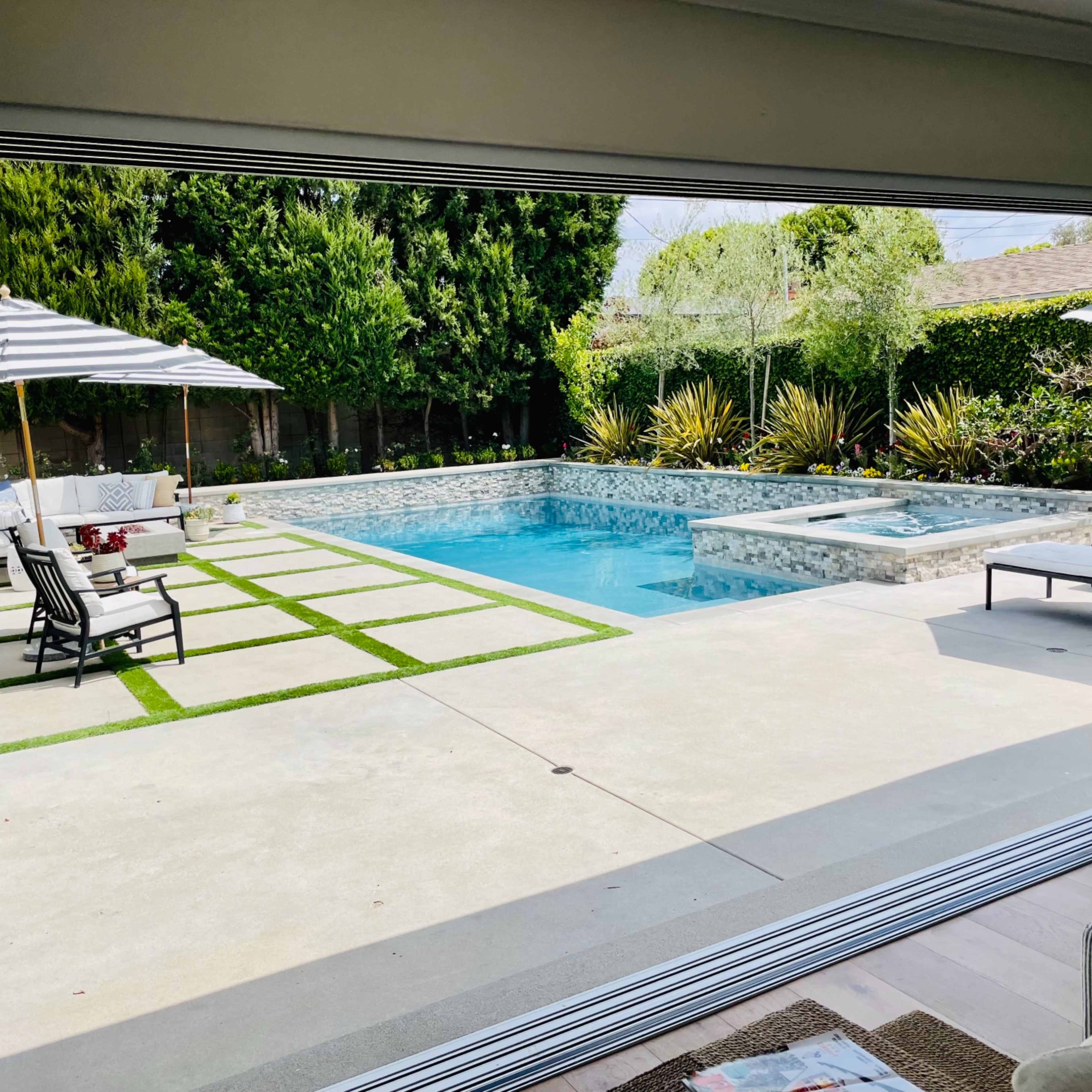 A view of a modern backyard featuring a swimming pool, spa, and lounge area with outdoor seating and an umbrella.