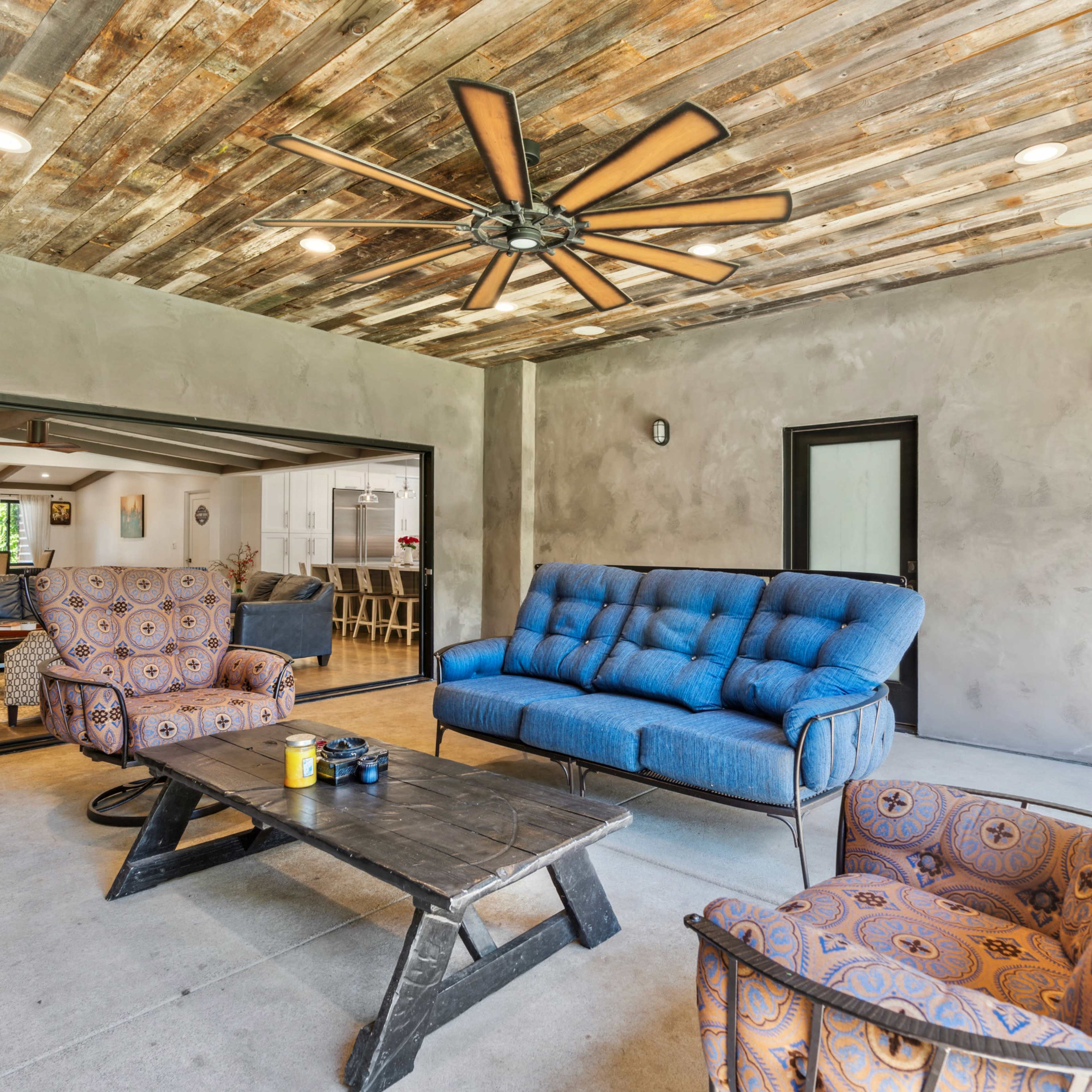 A cozy outdoor living space with a blue sofa and two patterned chairs arranged around a black wooden coffee table under a ceiling fan.