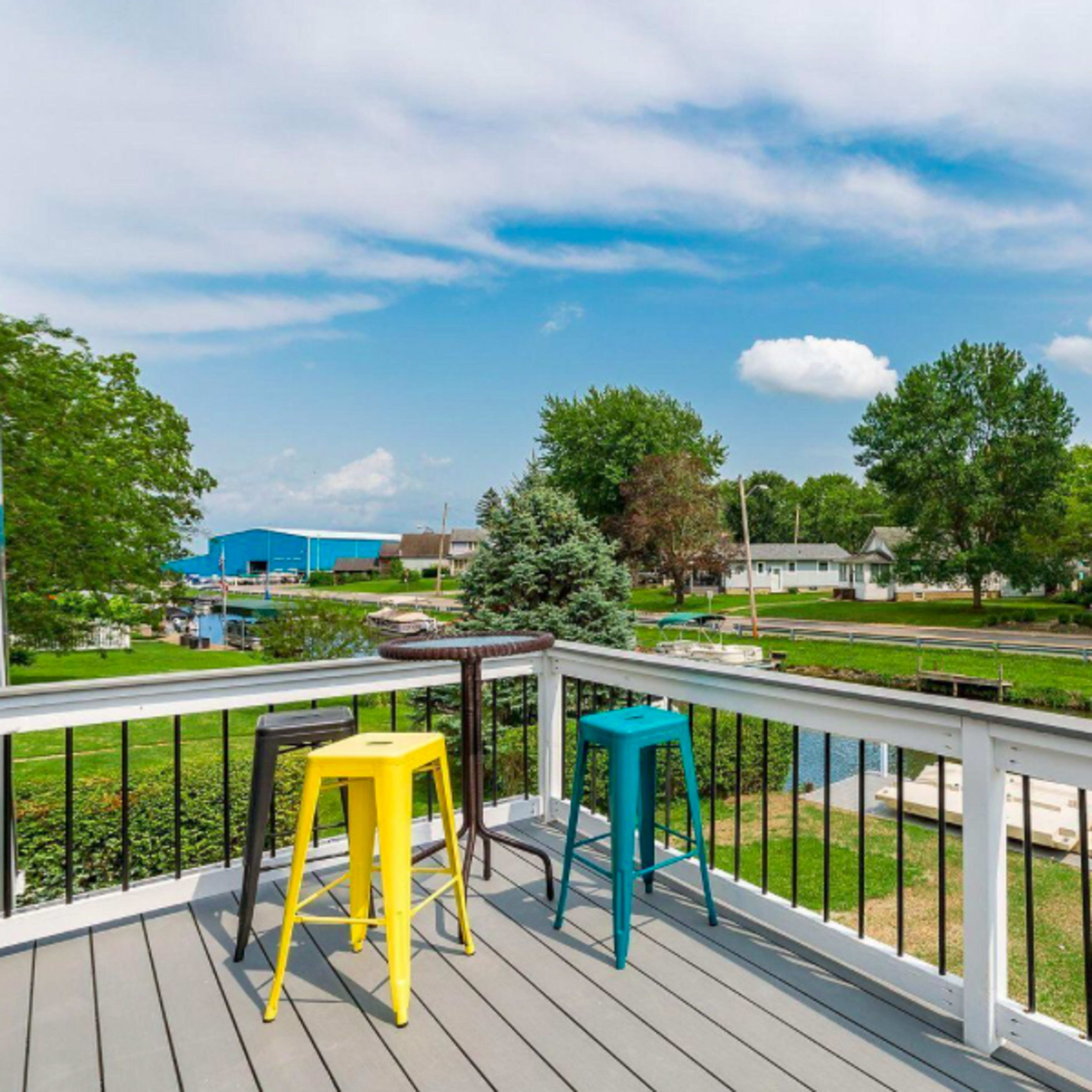 A deck features brightly colored stools overlooking a waterway lined with homes and trees under a partly cloudy sky.