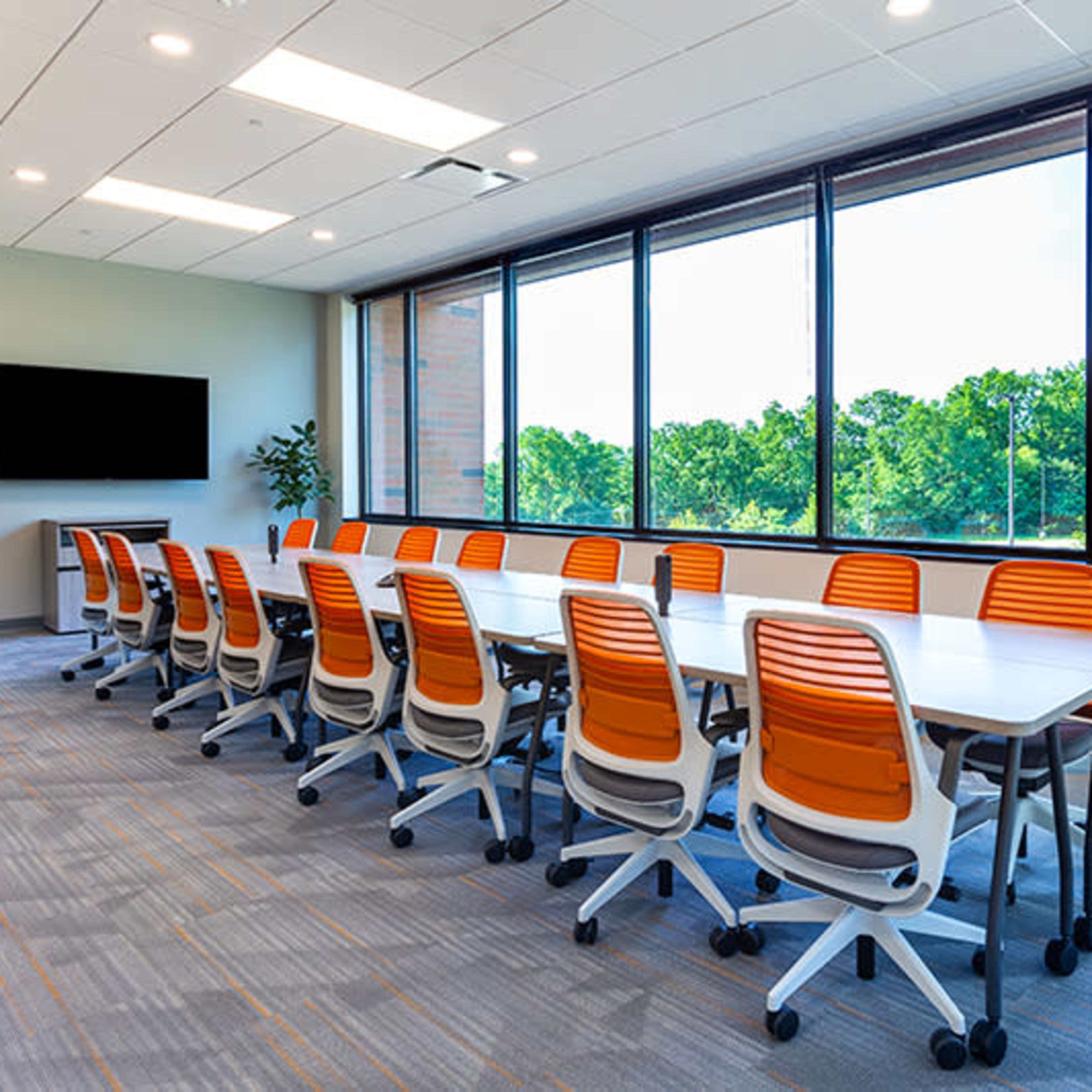 A large conference room with a long table and orange wheeled chairs is illuminated by natural light streaming through large windows overlooking green trees.