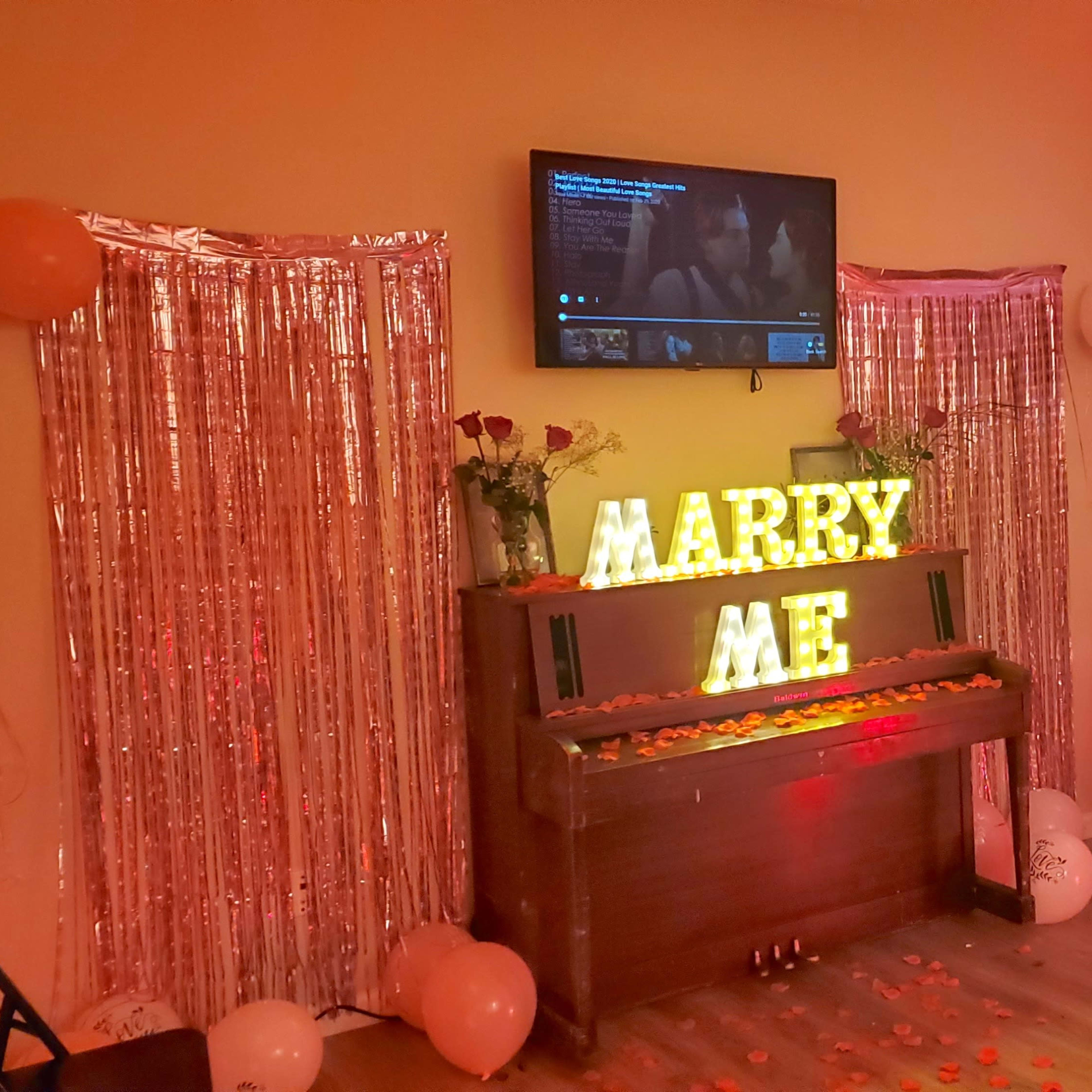 The scene features a small room decorated with orange lighting, a TV playing a video, a piano adorned with illuminated letters spelling "MARRY ME," and a table set with decorations for a celebration.