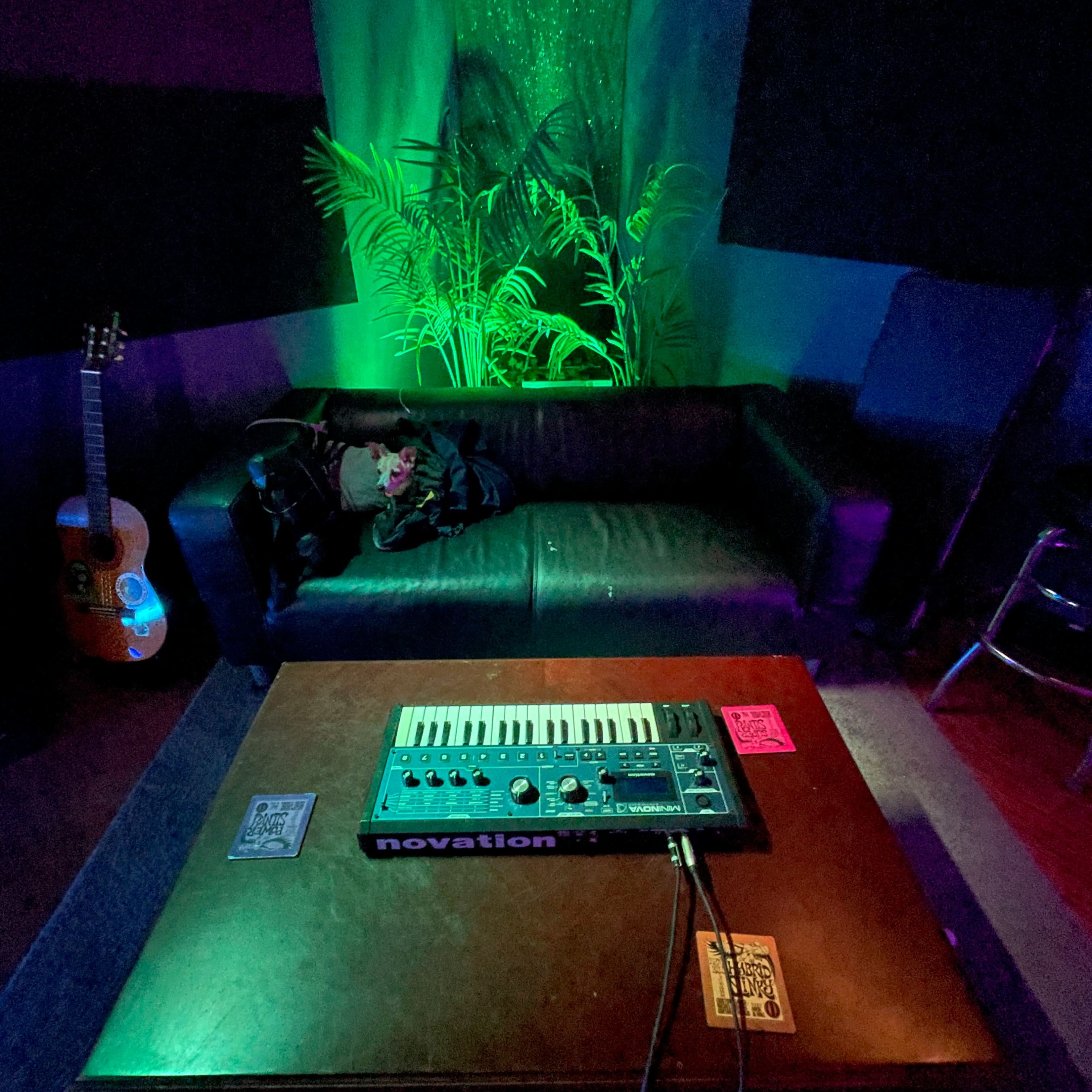 A dimly lit music studio with a keyboard on a table, a black couch, an acoustic guitar, and green lighting illuminating the space.