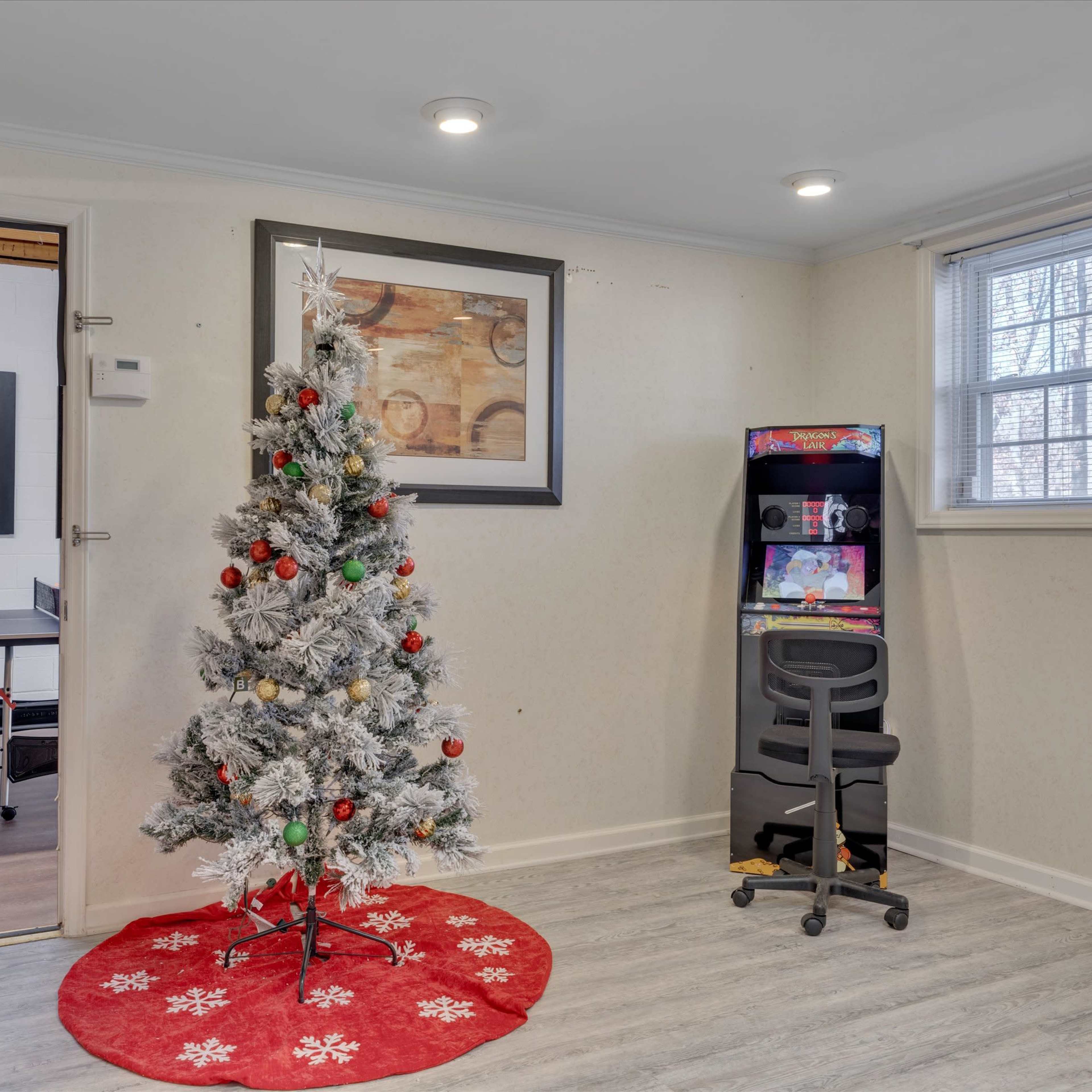 A decorated Christmas tree is positioned on a red circular rug in a room with a wall-mounted artwork and a small arcade machine beside a chair.
