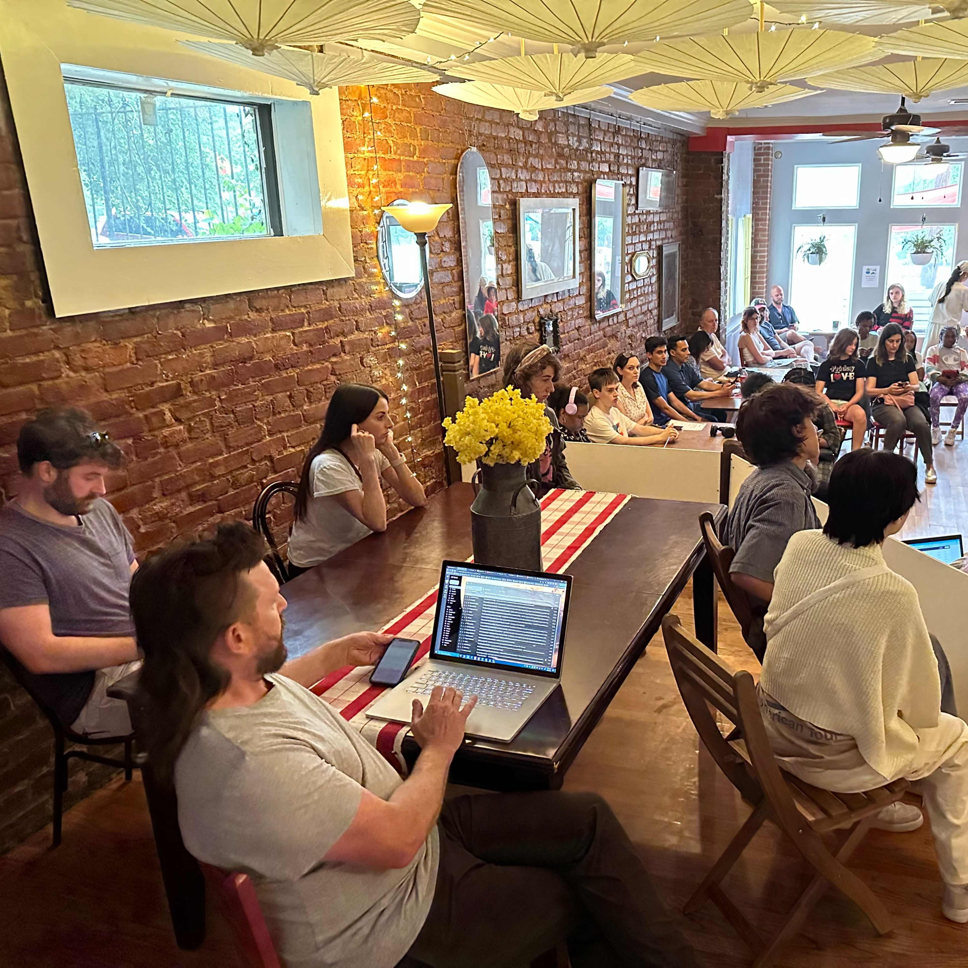 A group of people are gathered in a cozy room with exposed brick walls for a discussion or presentation, while others sit attentively in chairs.