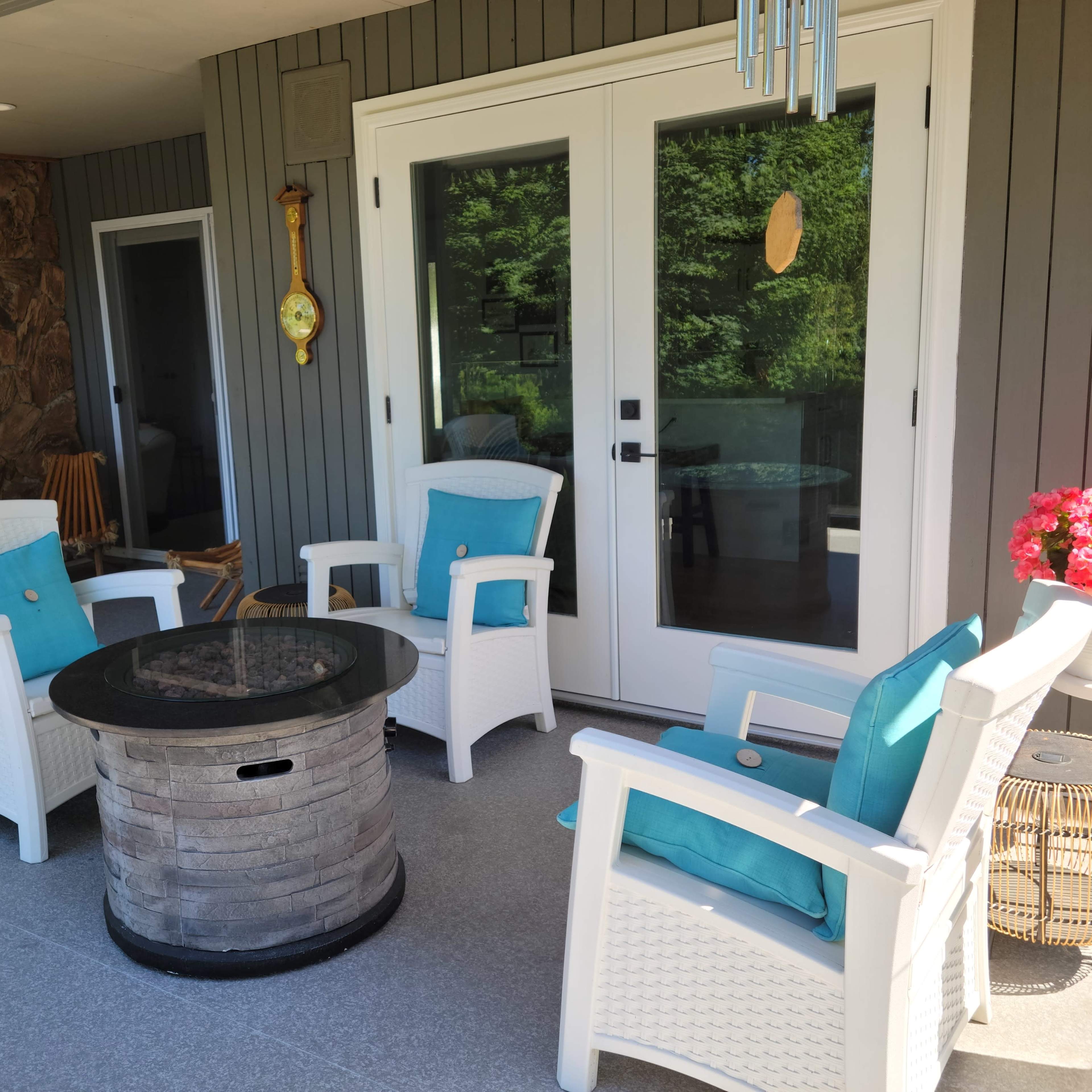 A outdoor seating area featuring four white chairs with turquoise cushions arranged around a circular table on a patio, with a stone wall and a barbecue grill in the background.