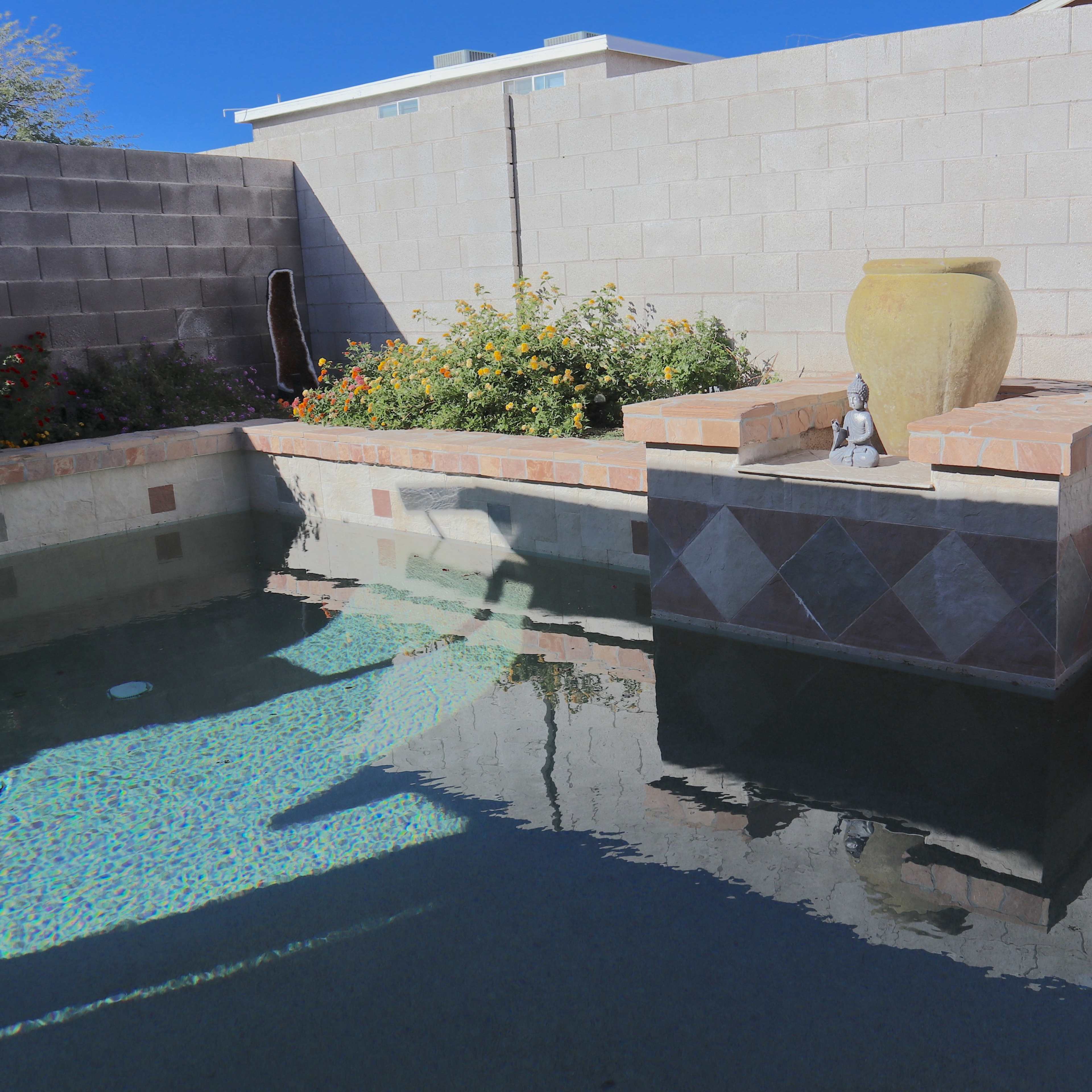A small, tranquil backyard pool area with a stone wall, potted plants, and a decorative water feature.