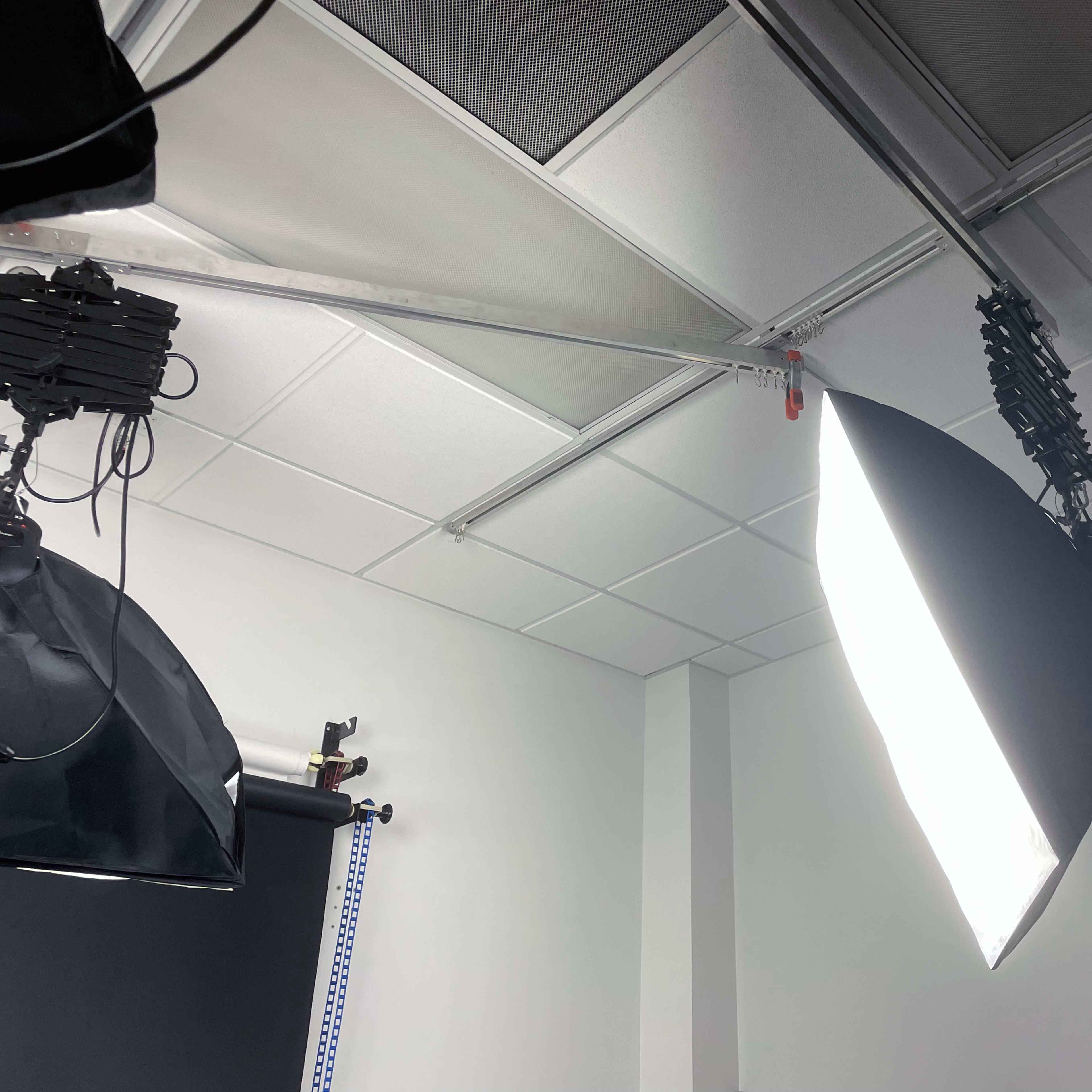 A photography studio setup with softbox lights mounted on the ceiling and a plain white wall in the background.
