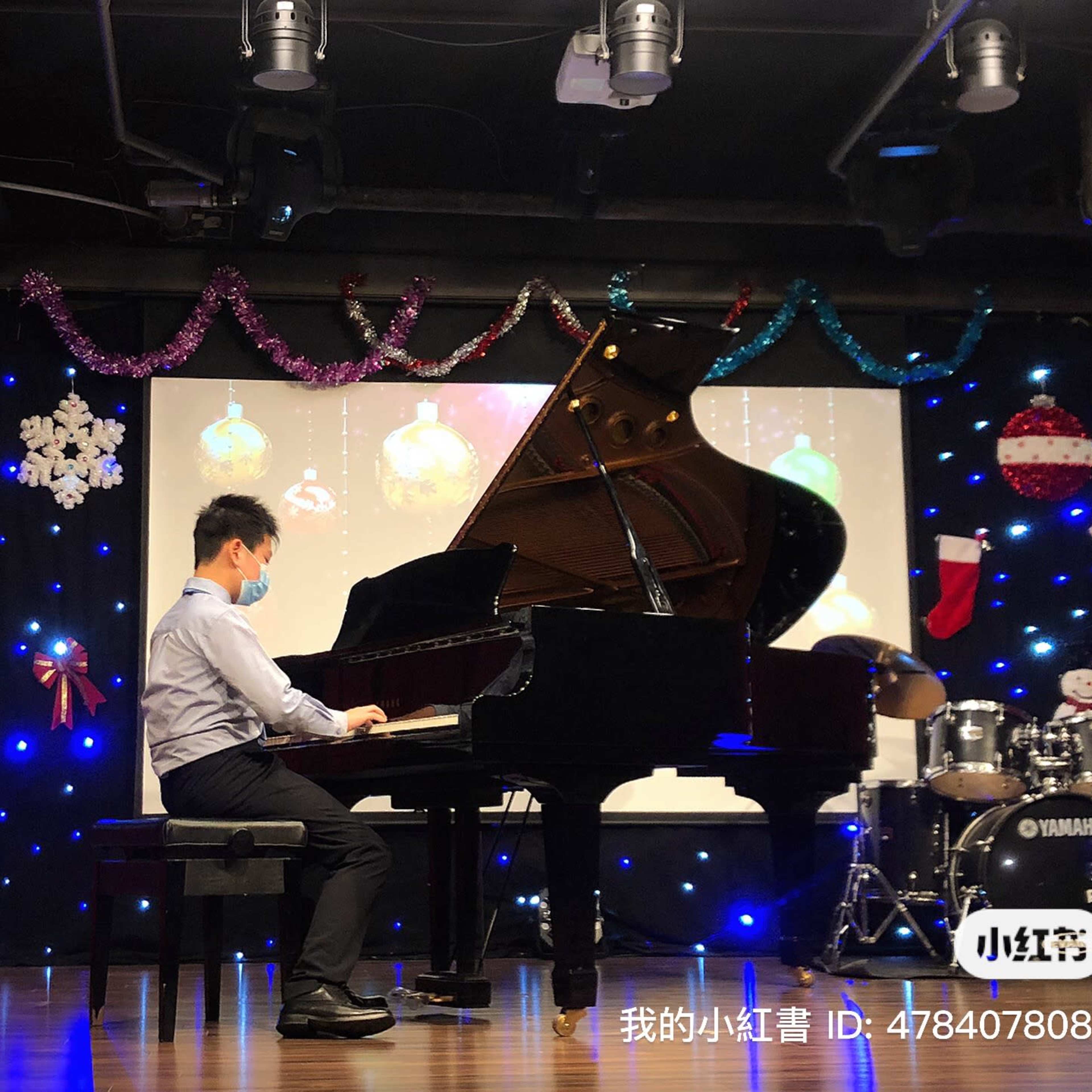 A person in a mask plays a grand piano on stage with decorative lights and a festive backdrop.