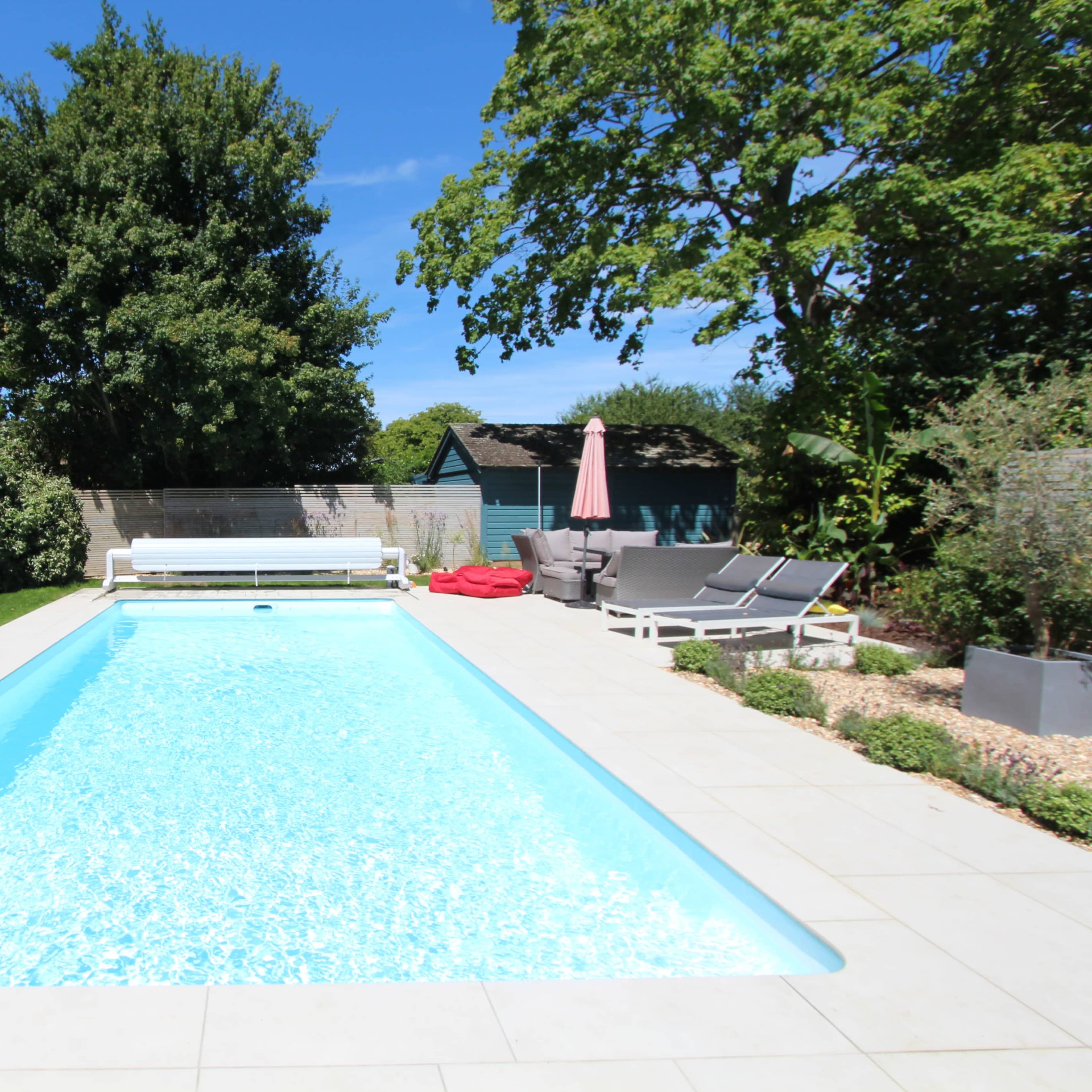 A swimming pool surrounded by a patio area with lounge chairs and a shaded seating space in a garden setting.