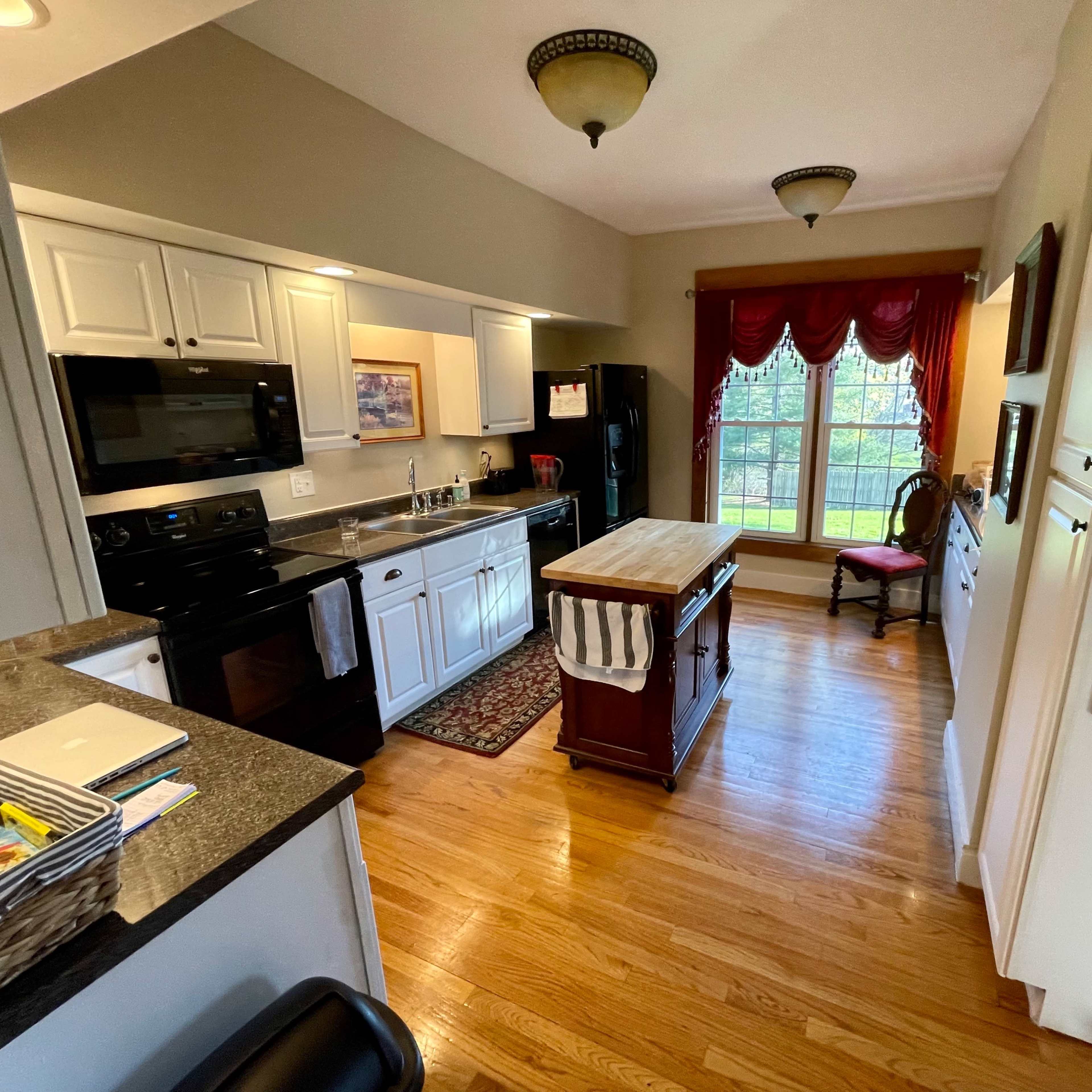 A kitchen features white cabinetry, black appliances, a central island, and a window with red drapes that overlooks a seated area.