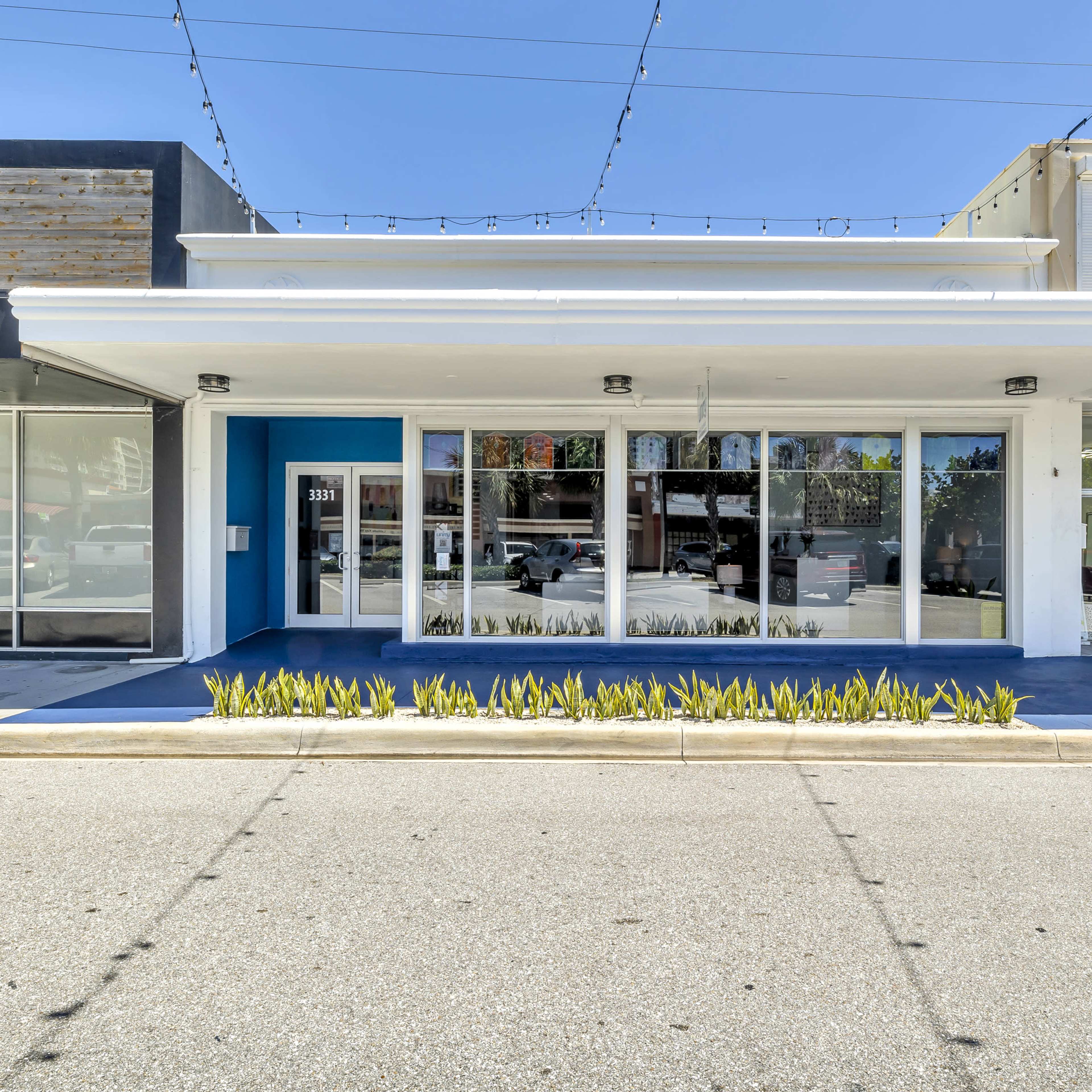 A shopfront with large glass windows and a blue accent wall, bordered by a tidy planter filled with greenery.