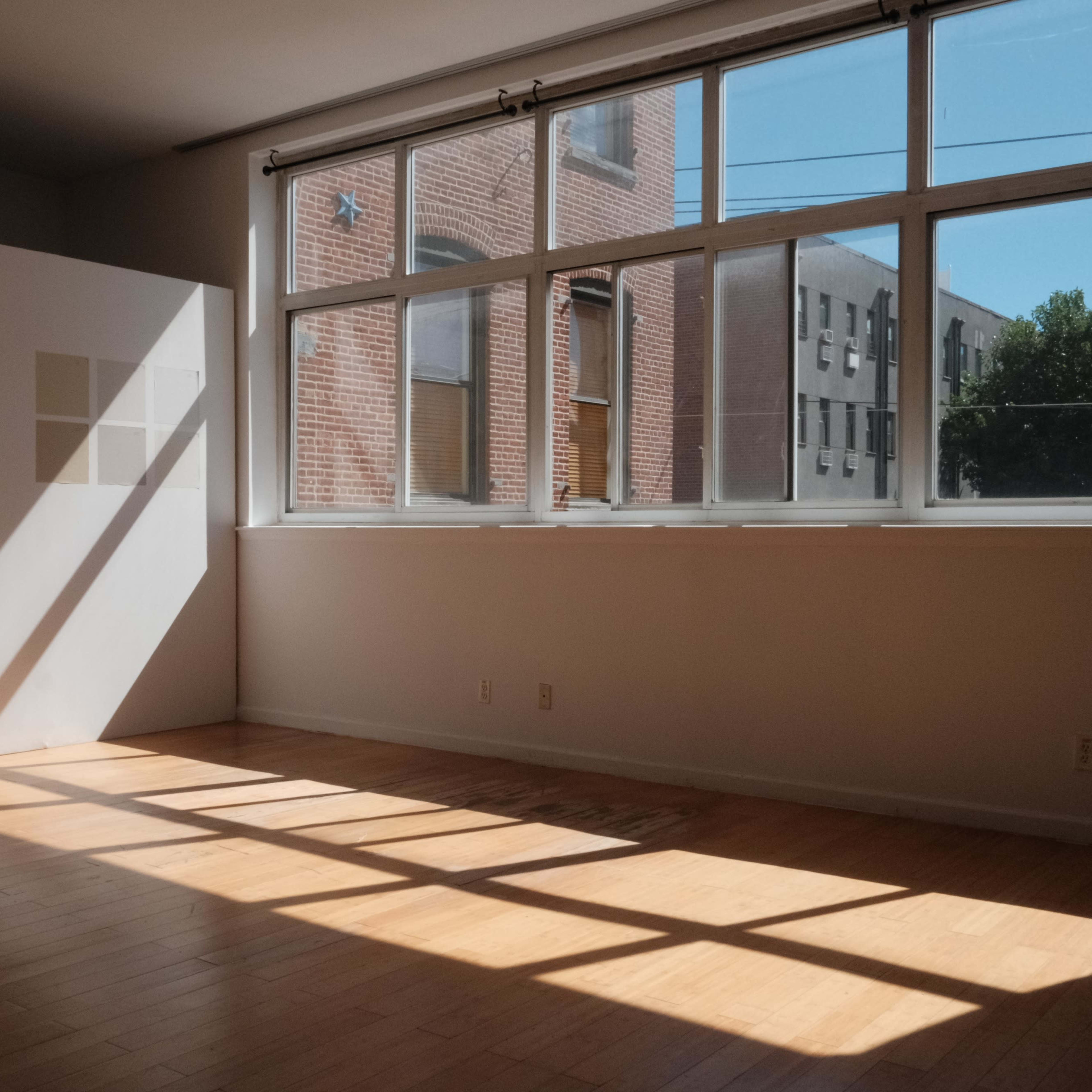 A sunlit room with wooden flooring and large windows casting shadows on a wall.